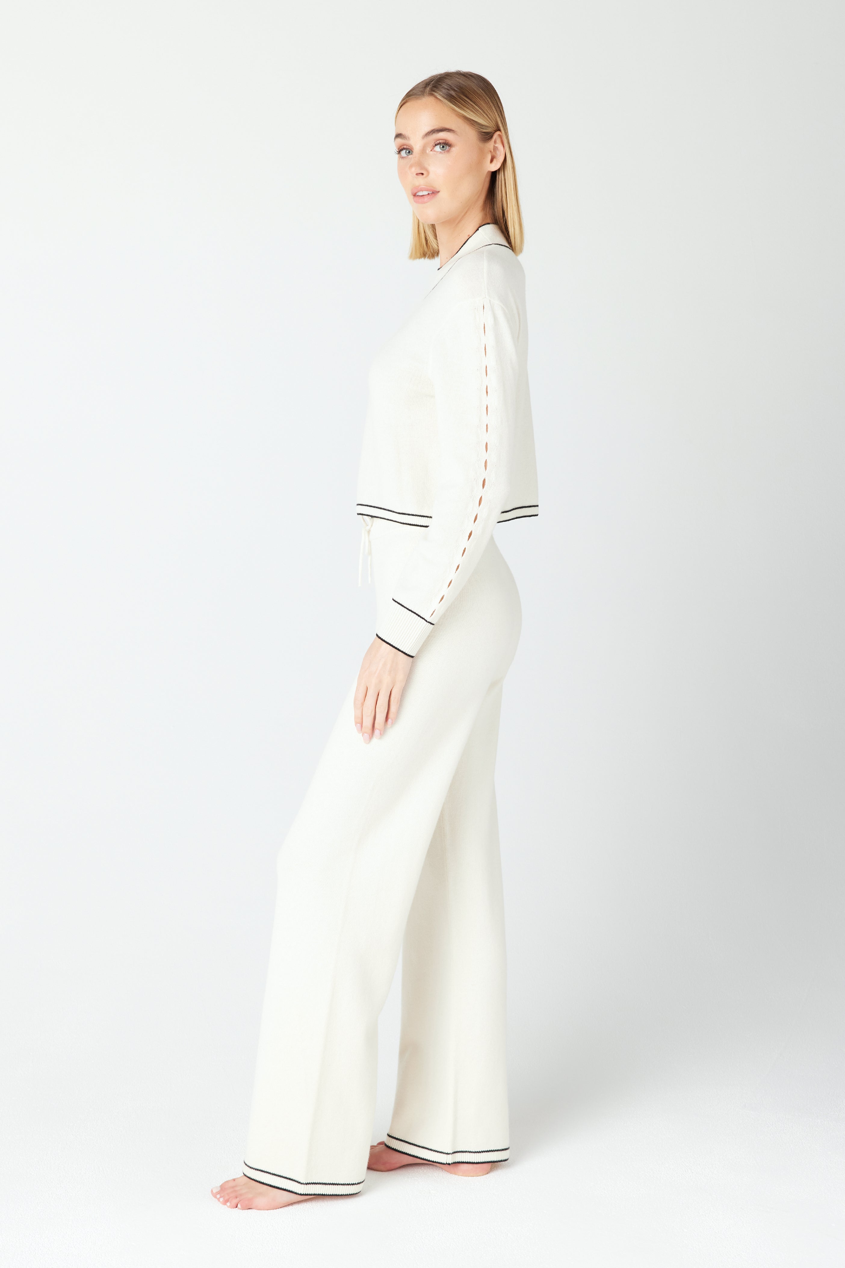 A woman exudes elegance in Gigi C Lounge loungewear, comprising a luxe long-sleeved top and Aubrie Wide Leg Pant with black trim. Her white ensemble harmonizes with the serene background as she gazes thoughtfully to her left, barefoot and effortlessly poised.