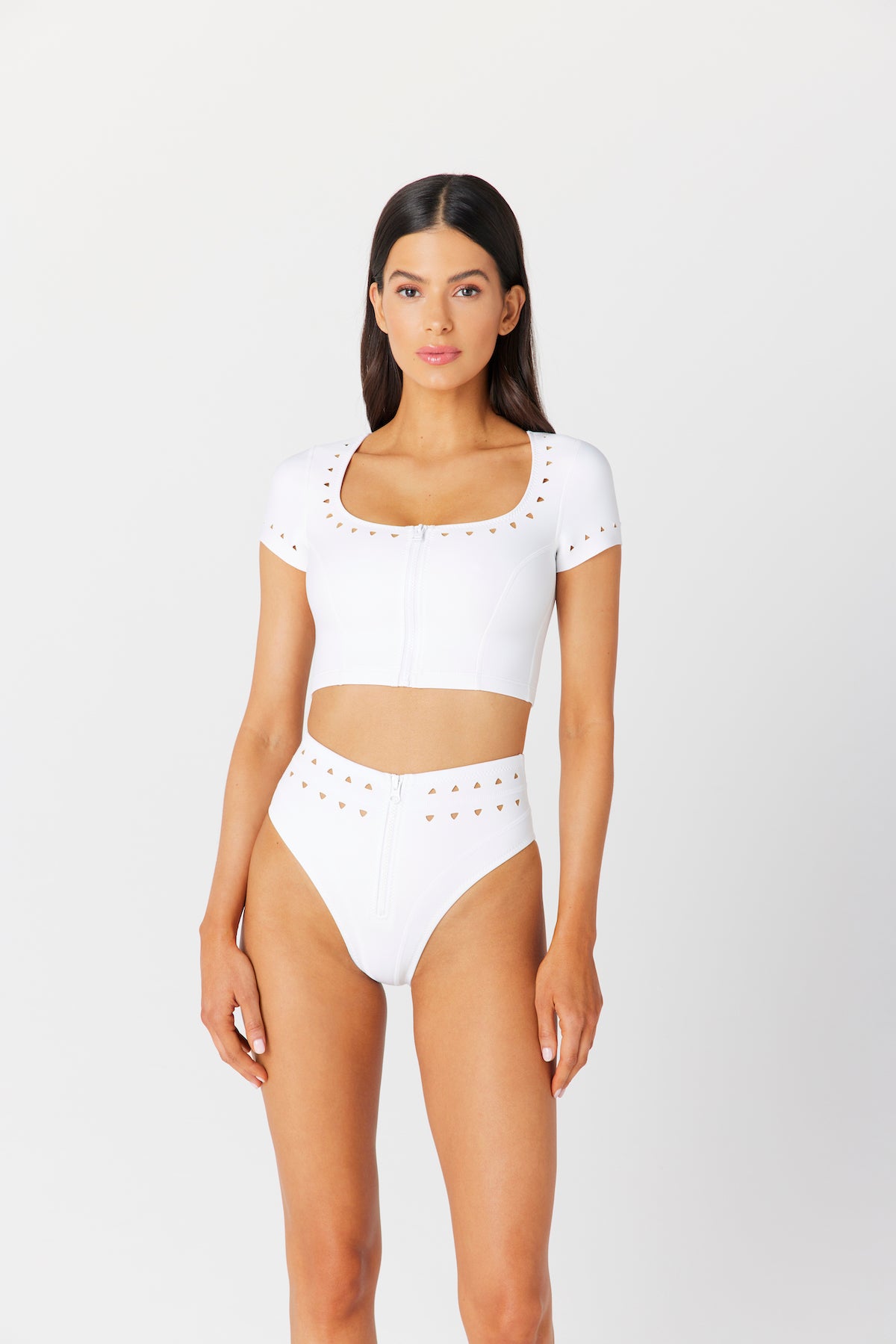 A woman in a stylish white Jennifer Crop Top with a decorative zipper by Gigi C Bikinis and high-waisted bottoms featuring scuba cutouts stands against a plain background, her long dark hair flowing as she confidently faces the camera.