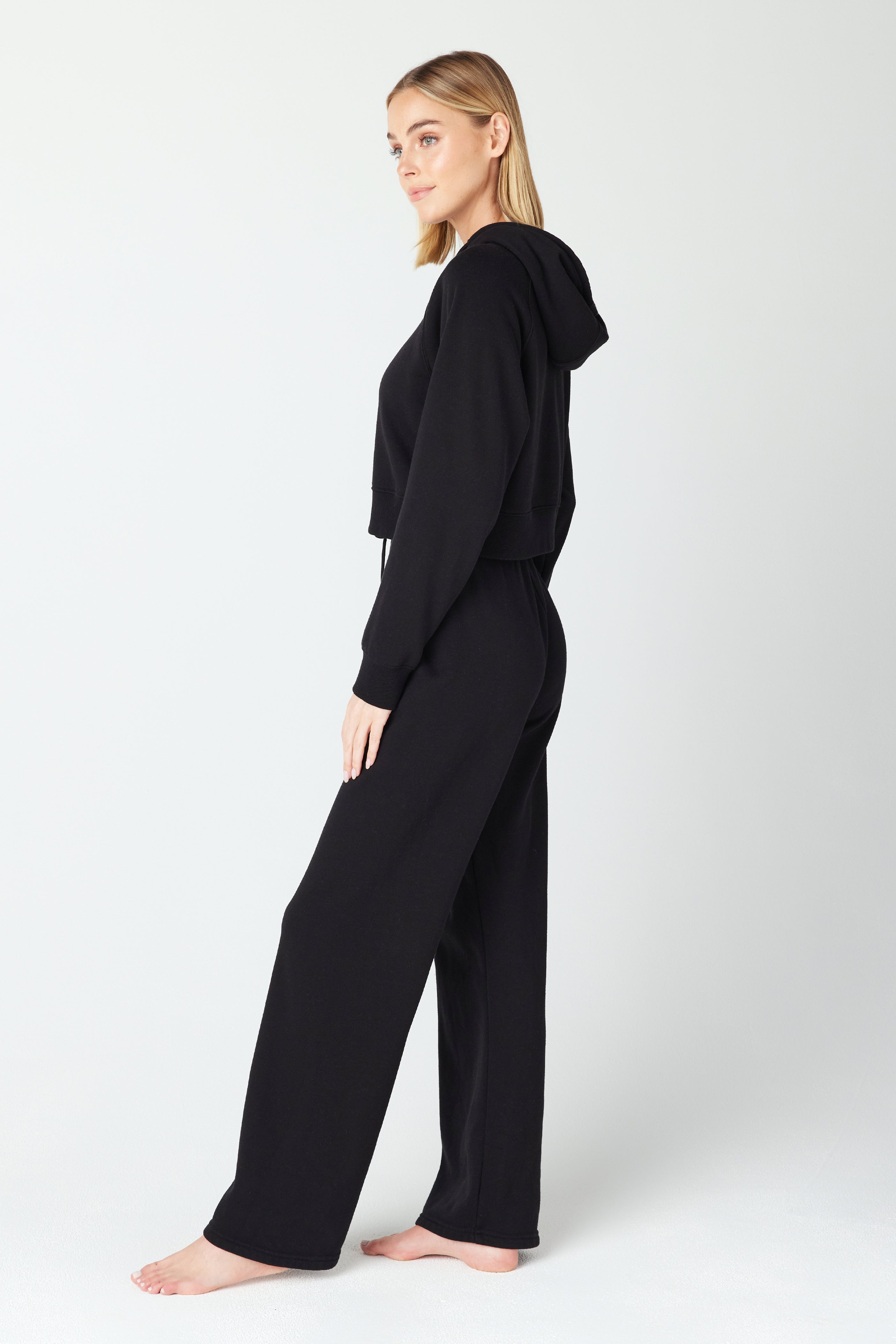 A person stands barefoot against a neutral background, wearing a black Juliet hoodie and Harlow Pant tracksuit from Gigi C Lounge. They face slightly right, looking ahead, showcasing the outfits long sleeves and relaxed pants for an effortlessly stylish look.