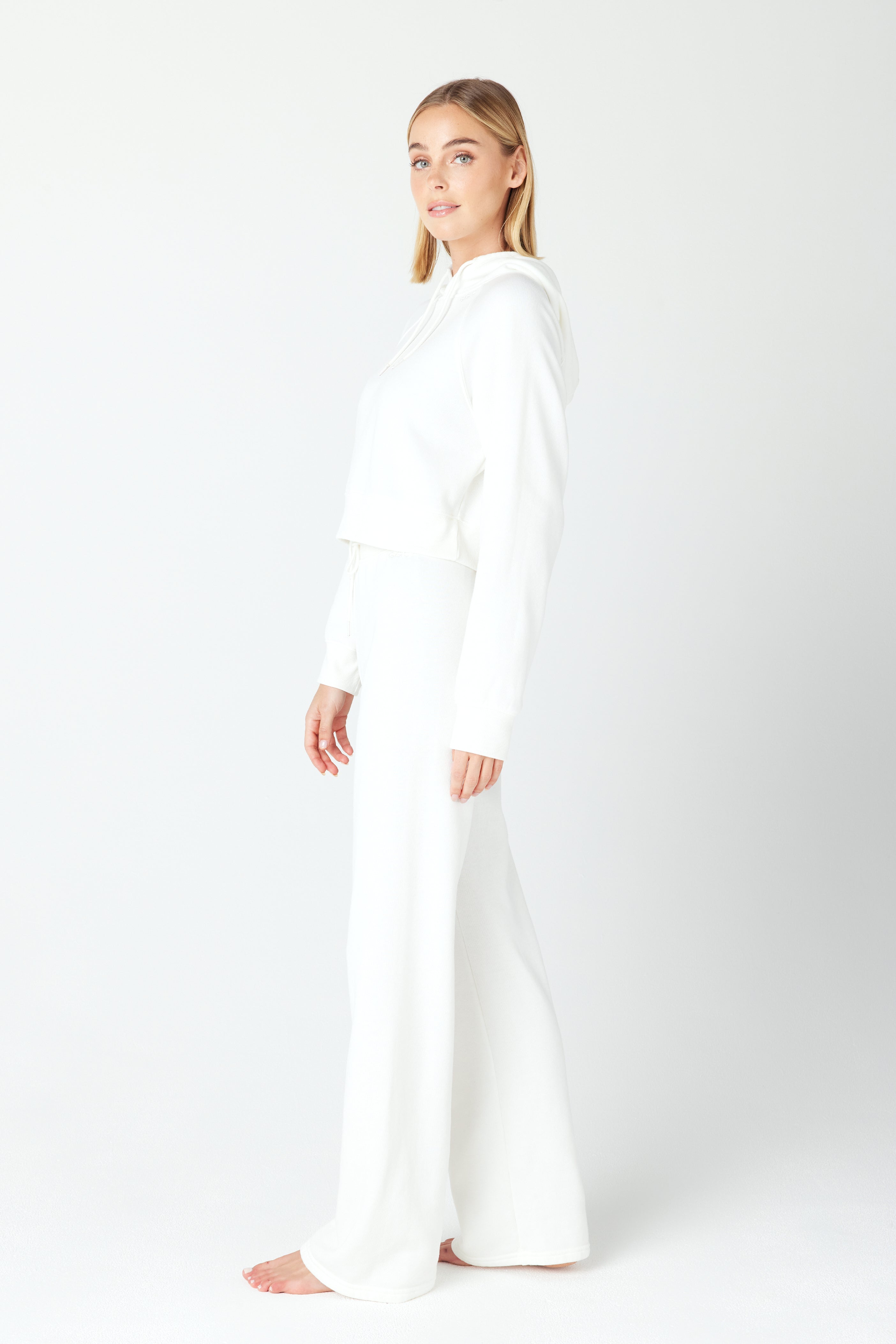 Against a white background, a person stands barefoot wearing the Juliet Hoodie by Gigi C Lounge, featuring an embroidered loop terry fleece and wide-leg pants. They gaze neutrally at the camera, hands resting naturally by their sides.