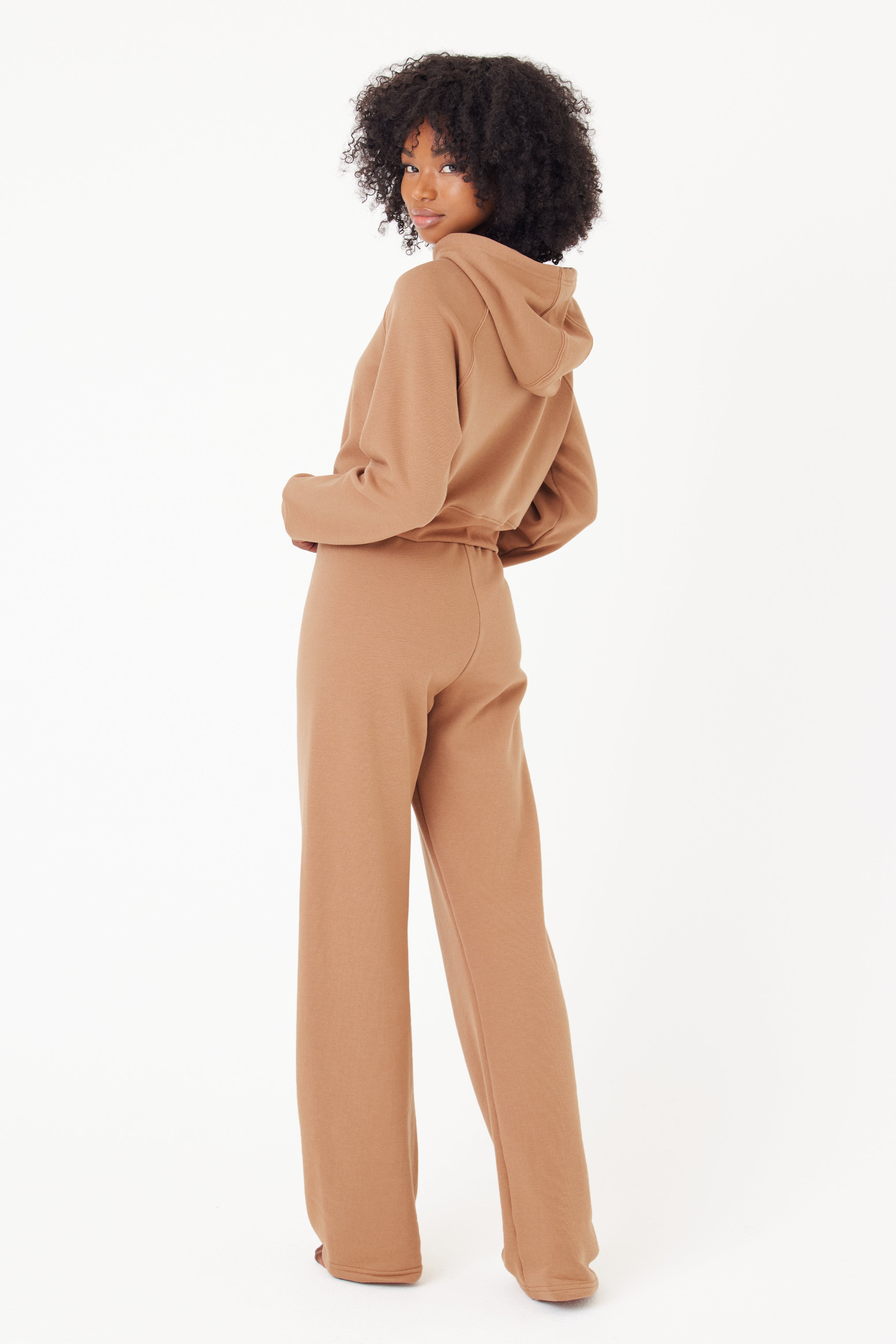 A curly-haired person models the light brown Juliet Hoodie and pants set by Gigi C Lounge, standing against a plain white background. The hoodie is subtly embroidered with GIGI C for an elegant touch as they glance over their shoulder.