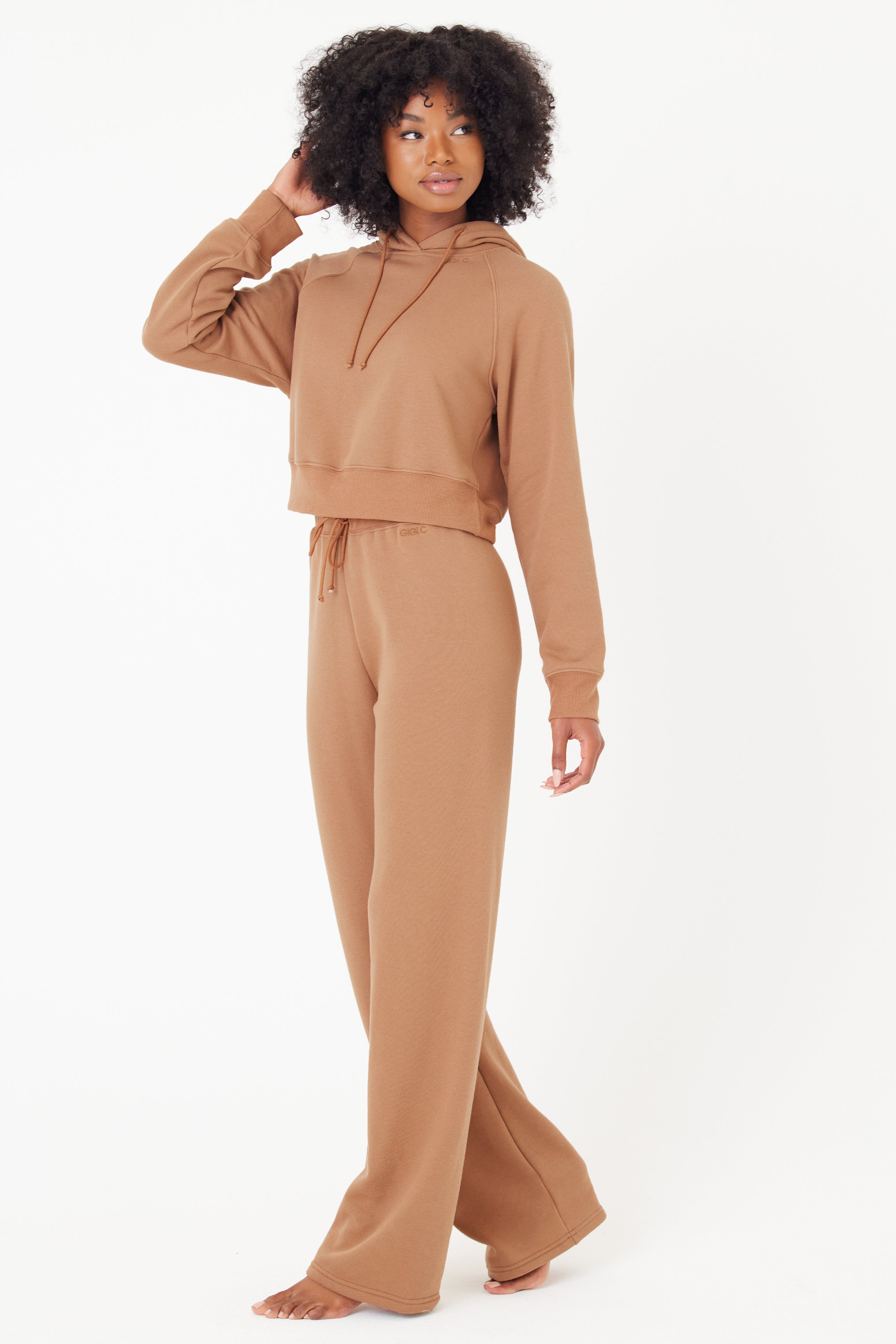 A curly-haired person is wearing a brown Juliet Hoodie and matching pants from Gigi C Lounge. The GIGI C embroidery adds elegance as they pose barefoot, with one hand in their hair, looking slightly to the side against a plain white background.