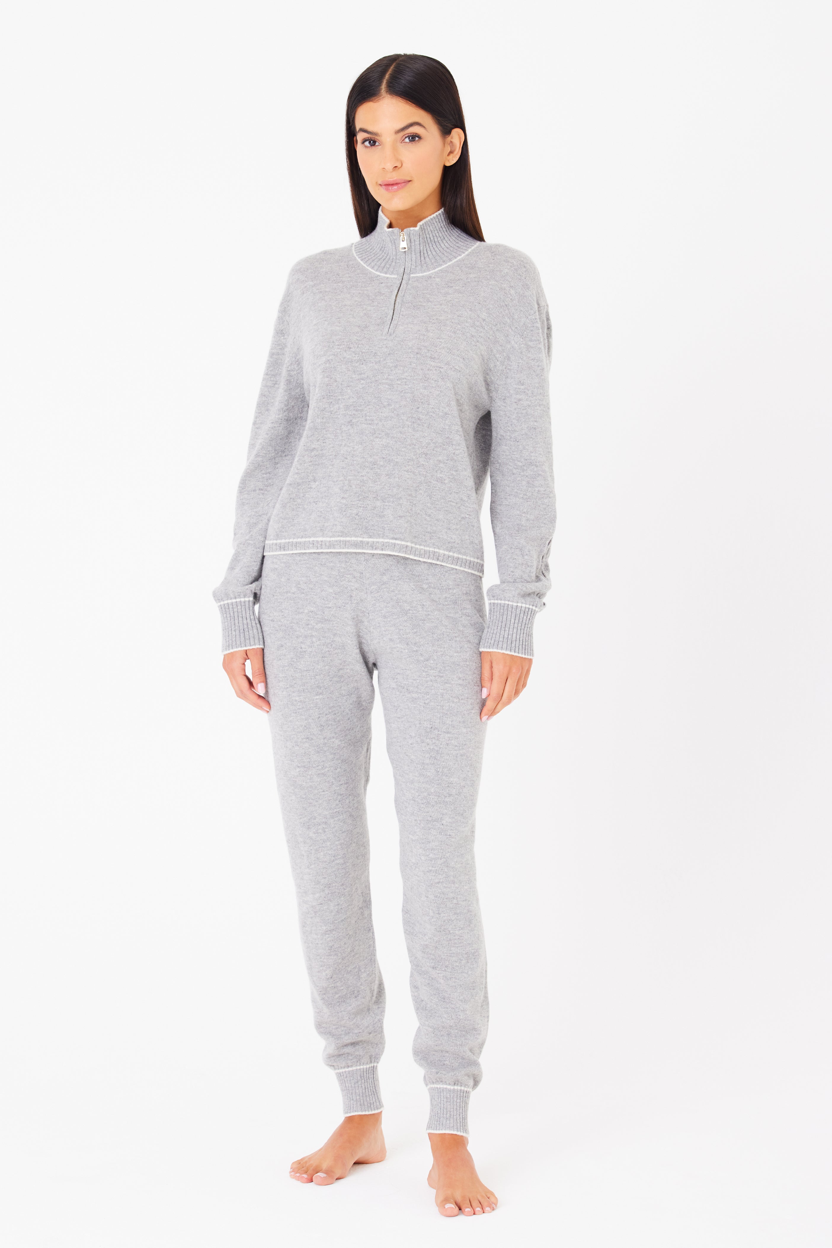 A person with long, dark hair stands barefoot against a white backdrop, wearing the Gigi C Lounges Alice Jogger set in gray. Made from a cozy wool/cashmere blend, it features a long-sleeve pullover and matching joggers, creating a comfortable and casual look. They have a neutral expression.