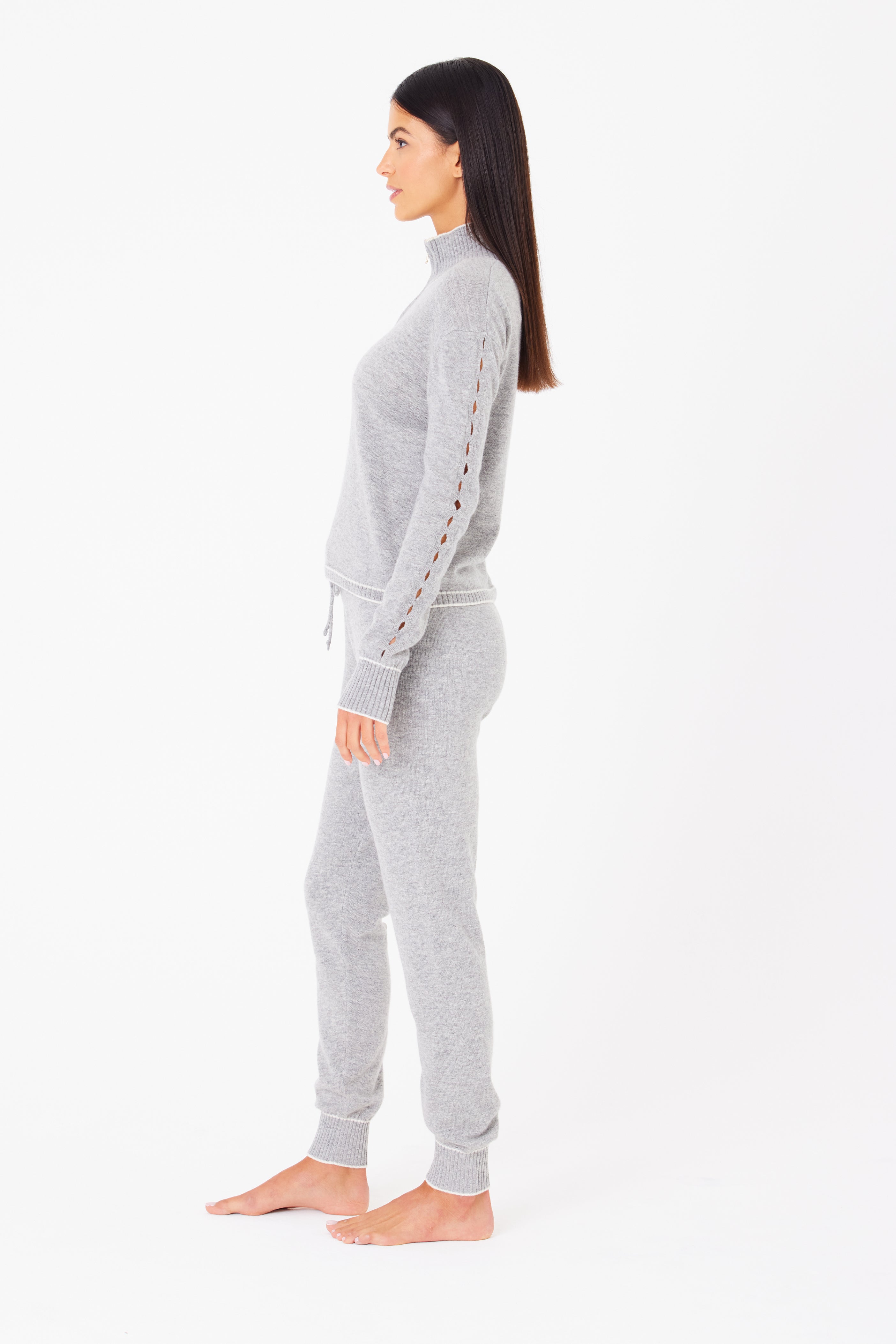 A woman with long dark hair stands barefoot in profile against a white backdrop, wearing Gigi C Lounges light gray knit sweater made from a luxurious wool/cashmere blend and matching Alice Jogger pants, detailed with buttons along the sleeves.