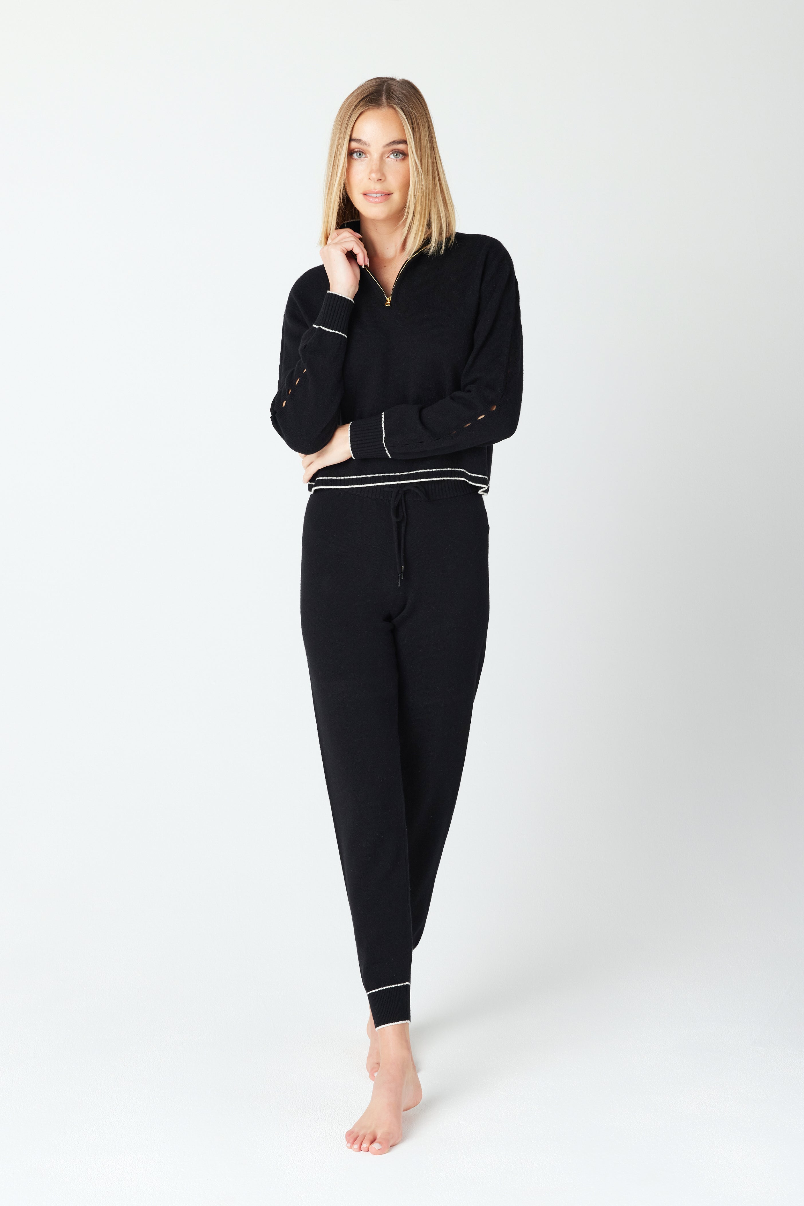 A person with long blond hair is wearing the black June Sweater featuring a Gigi C gold half zipper, paired with matching black pants with white trim. They stand barefoot against a plain white background, smiling while gently touching their face.