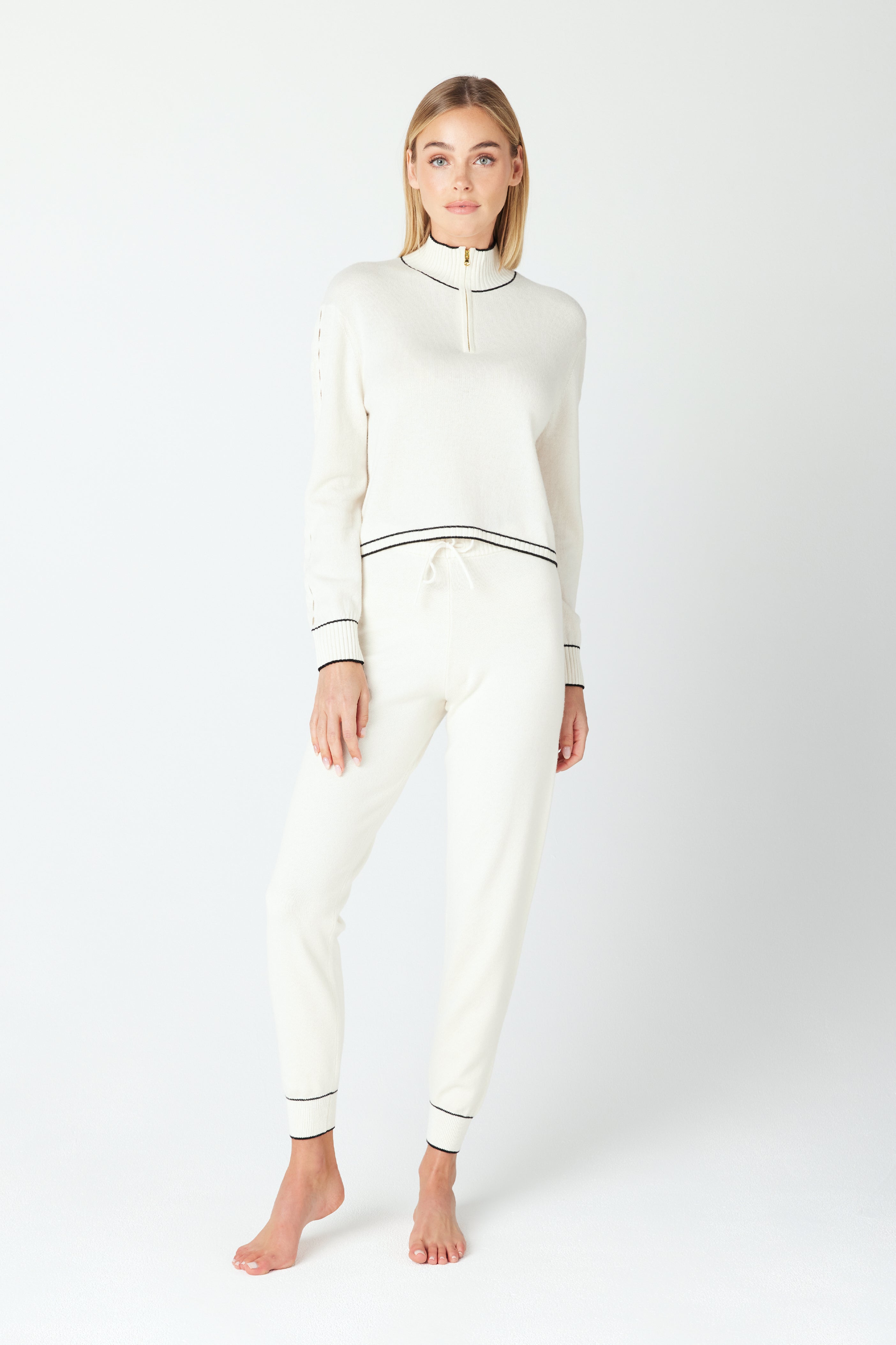 A person stands barefoot in a June Sweater by Gigi C Lounge, an off-white knit made from recycled cashmere, paired with pants featuring black trim. Their long hair cascades as they look at the camera against a plain light background.