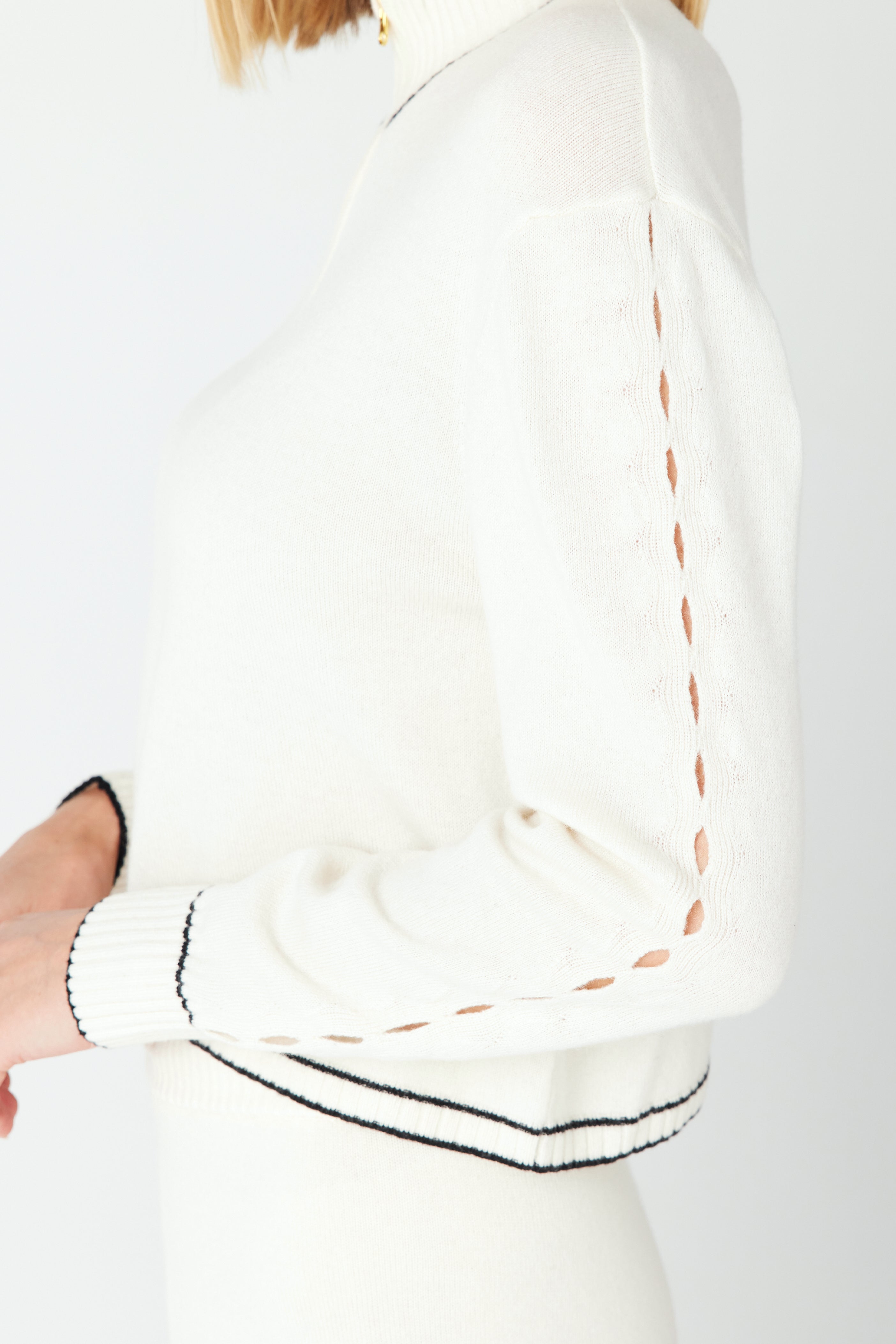 A person wears a Gigi C Lounge June Sweater made from recycled cashmere, with black trim and eyelet sleeve details, standing sideways with one hand on their hip against a white background.