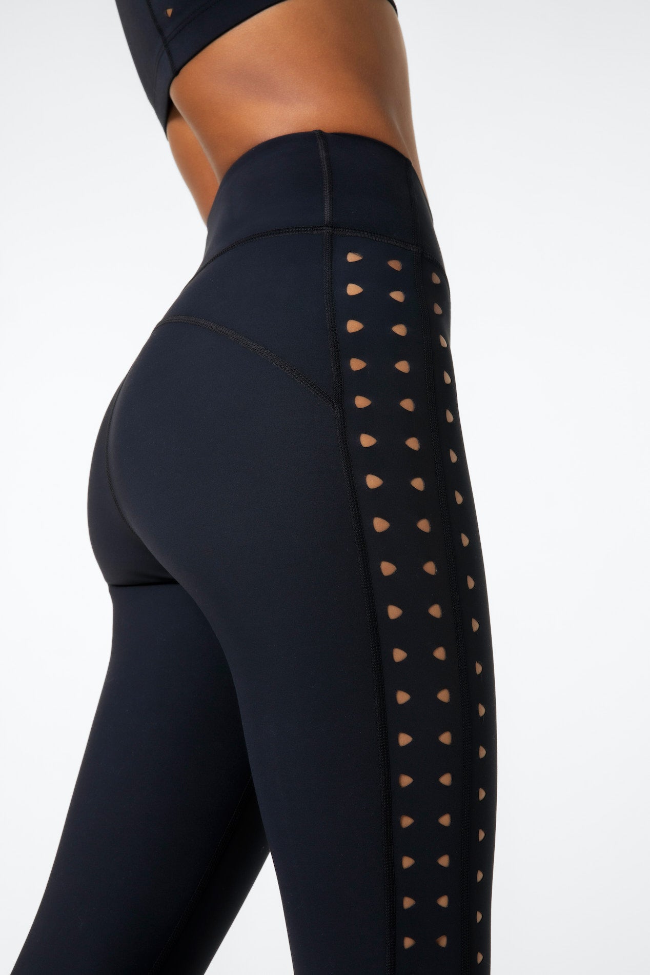 Close-up of someone in Gigi C Sports Katrina Legging, showcasing the black compression fabric with small laser cutouts along the side. The focus is on the lower torso and legs, highlighting the sleek design against a plain, light background.