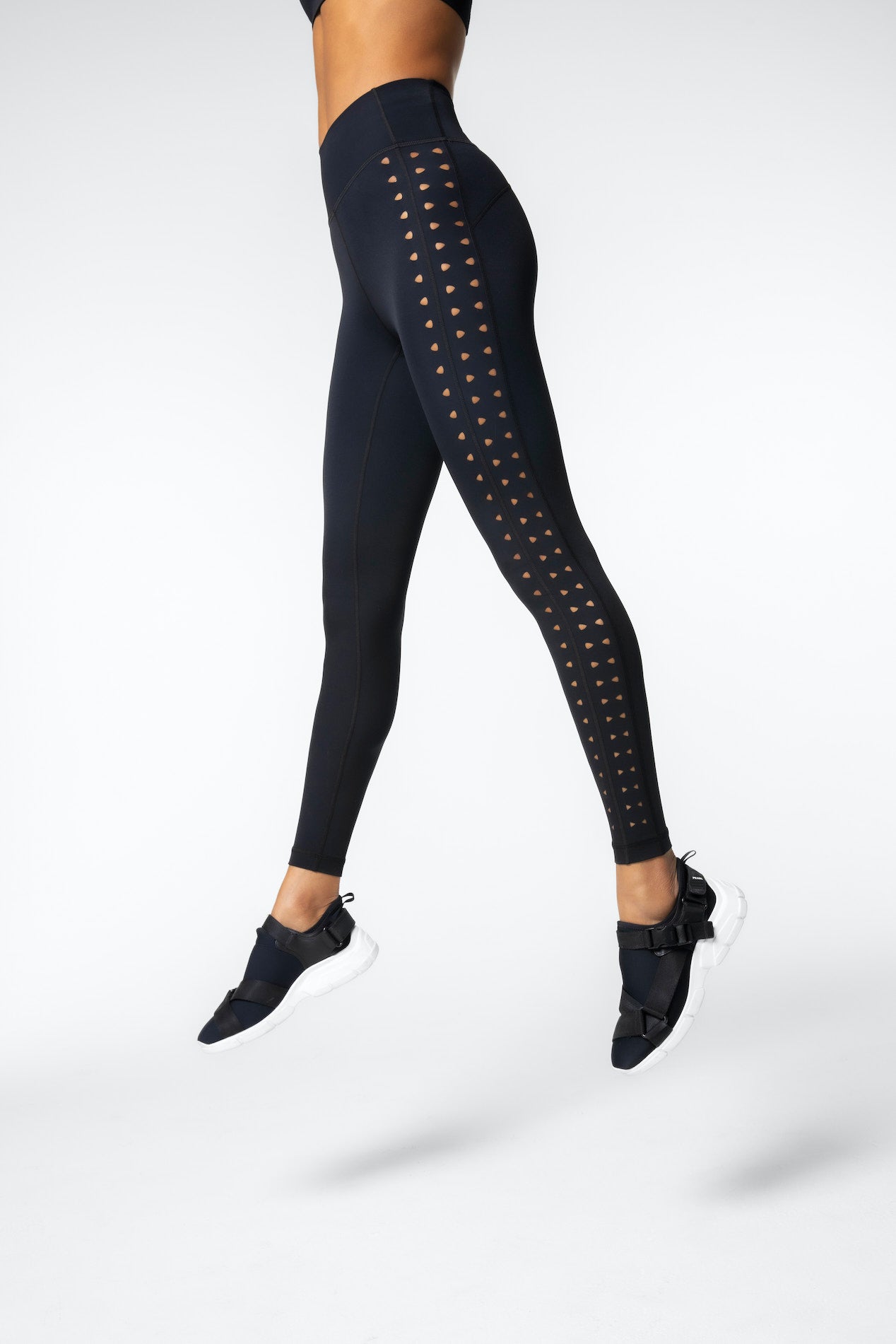 A person is mid-jump wearing Gigi C Sports stylish black high-waisted Katrina Legging with side laser cutouts, complemented by sleek black sneakers with white soles, all against a plain light gray backdrop.