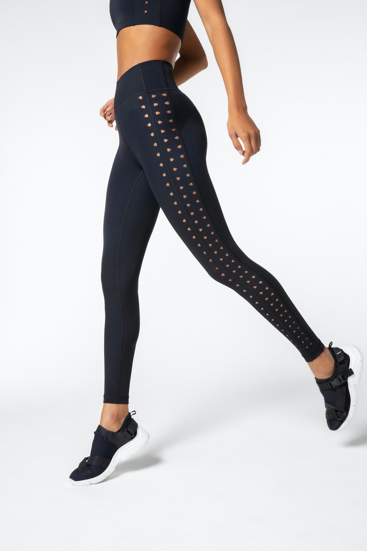 A person in black athletic wear is walking, highlighting a crop top and Gigi C Sports Katrina Legging, featuring stylish laser cutouts along the sides. Theyre in motion, lifting one leg while wearing black sneakers with white soles crafted from compression fabric for optimal support.