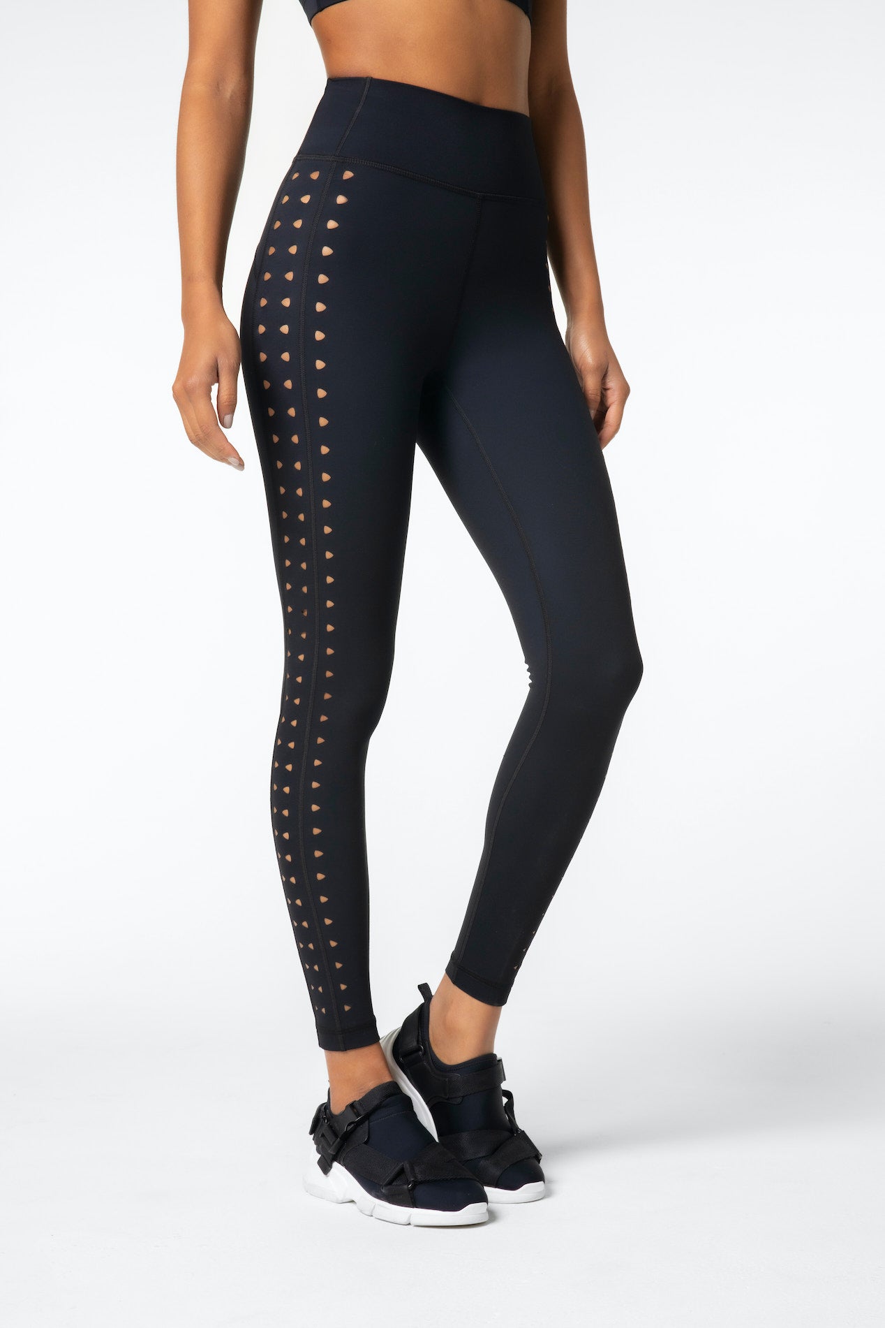 Someone wearing high-waisted Katrina Legging by Gigi C Sport in black, showcasing a chic laser cutout pattern on the sides. Paired with black sneakers, this striking look pops against a plain white background, emphasizing the stylish athletic wear.