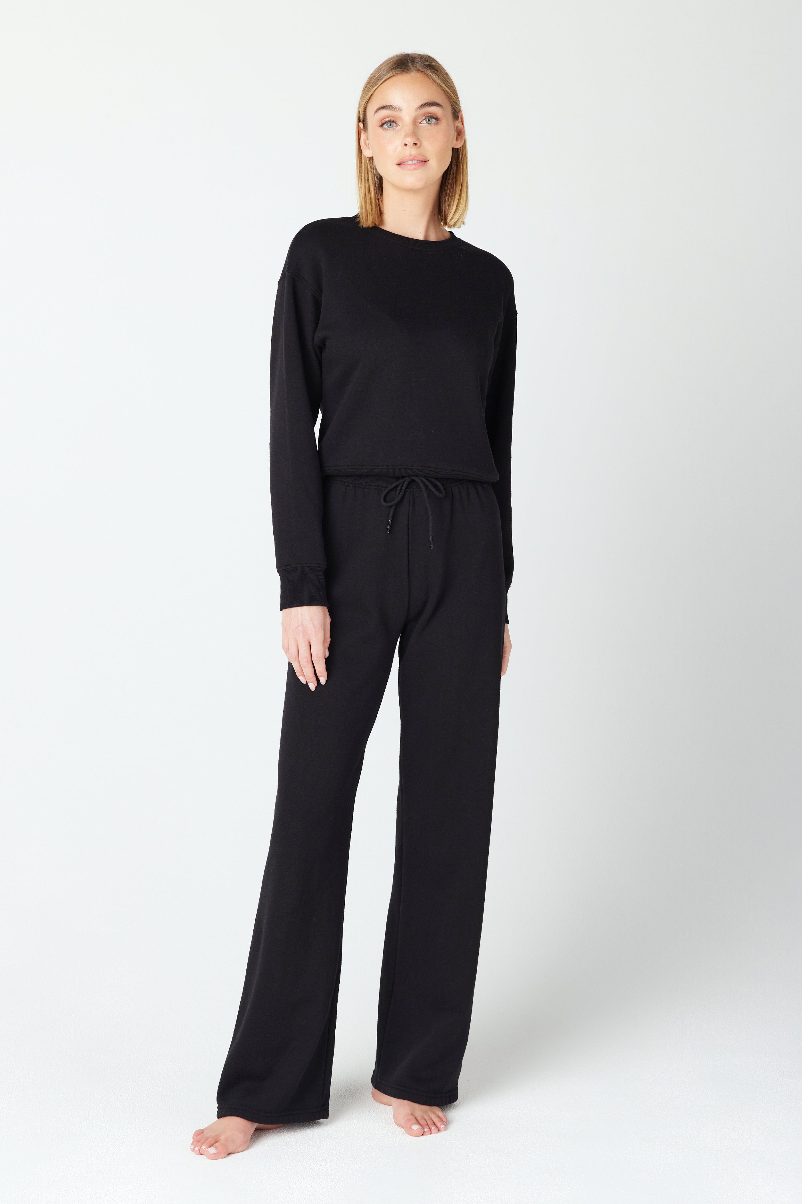 A person stands against a light background, wearing a black Kenzie Pullover by Gigi C Lounge and matching drawstring pants in soft Loop Terry Fleece. Barefoot, with their gaze slightly off-camera, they exude casual elegance.