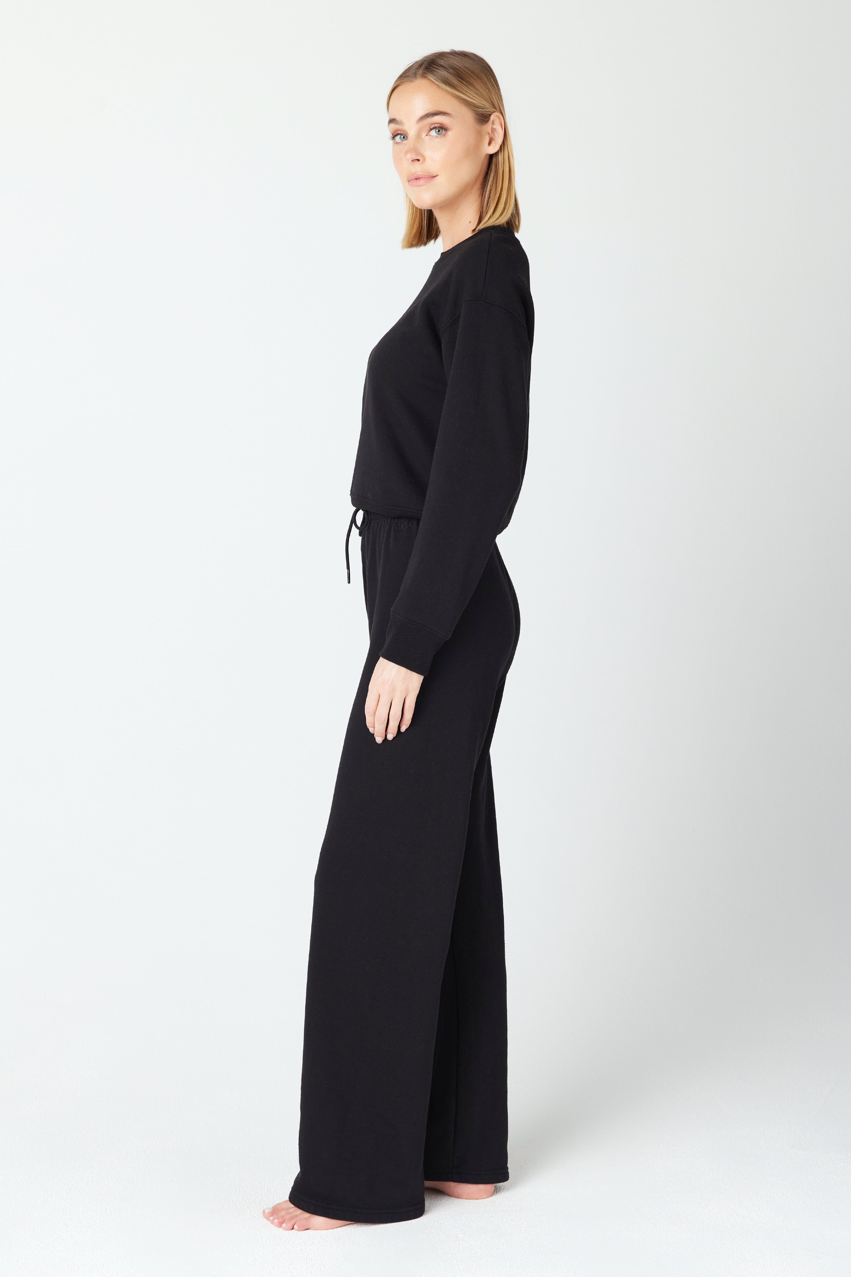 A person with shoulder-length blonde hair poses barefoot in a profile view, wearing a matching black Kenzie Pullover and wide-leg pants made from Loop Terry Fleece by Gigi C Lounge. The minimalist elegance of the ensemble is complemented by the plain, light-colored background.