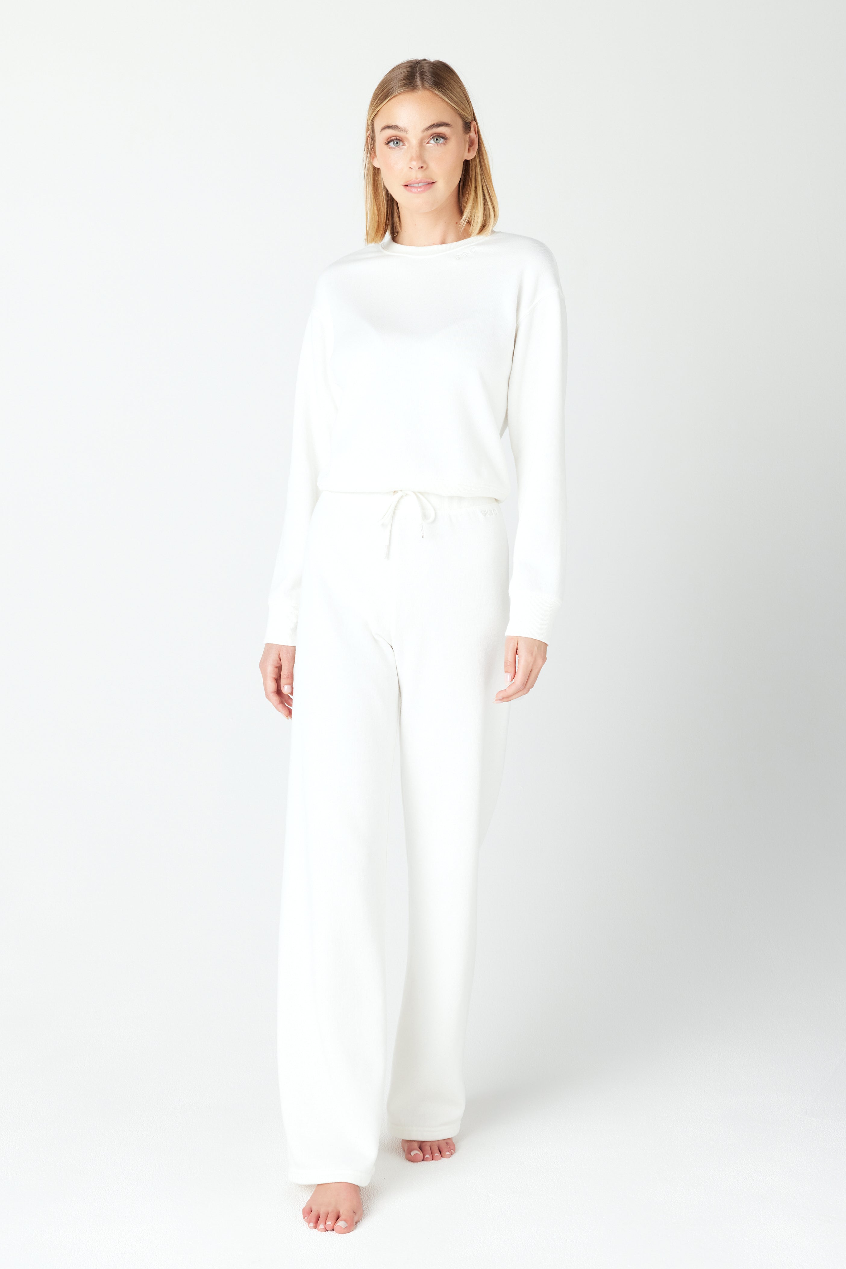 Against a plain backdrop, a barefoot individual with medium-length hair wears Gigi C Lounges white Kenzie Pullover and matching wide-leg pants in Loop Terry Fleece, creating a casual and relaxed style ideal for leisurely moments.