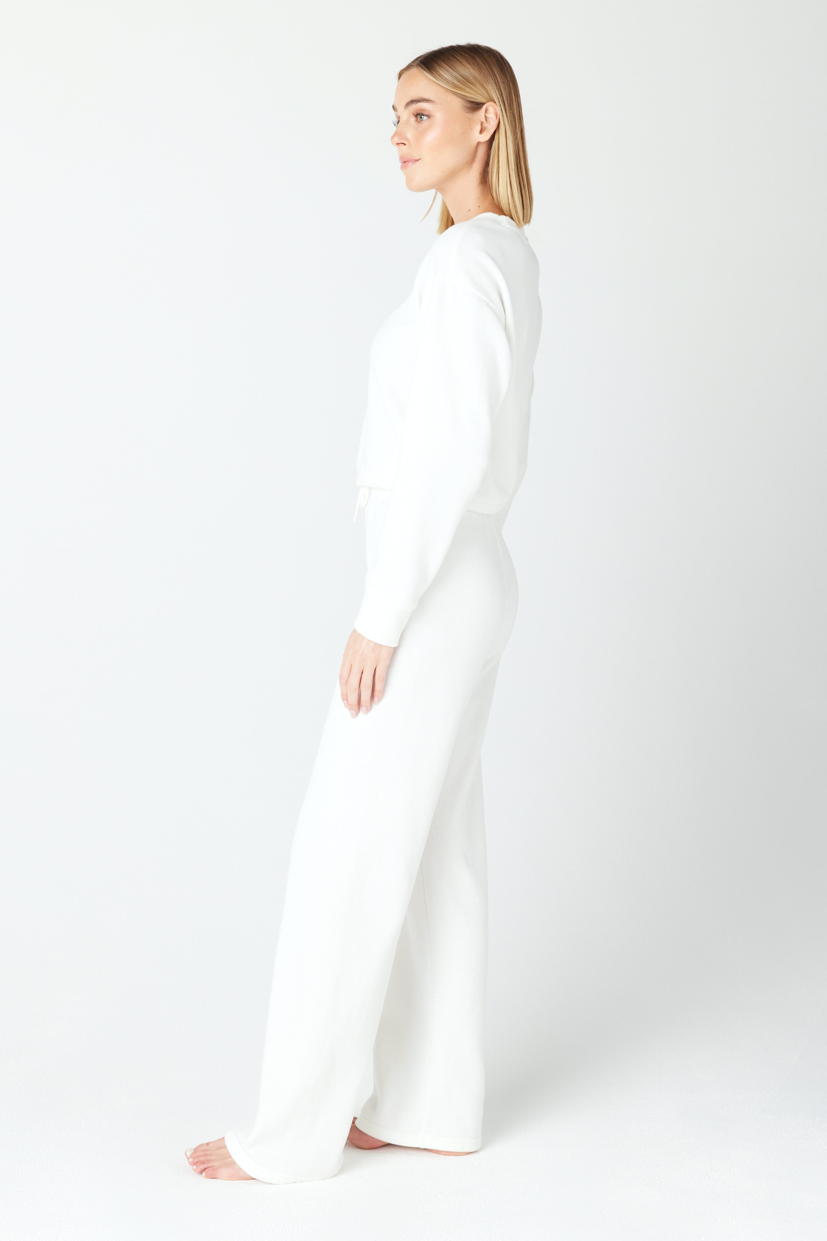 A person stands on a white background wearing a Gigi C Lounge Kenzie Pullover paired with wide-leg pants. Barefoot and facing left, their arms are relaxed by their side, exuding effortless elegance.