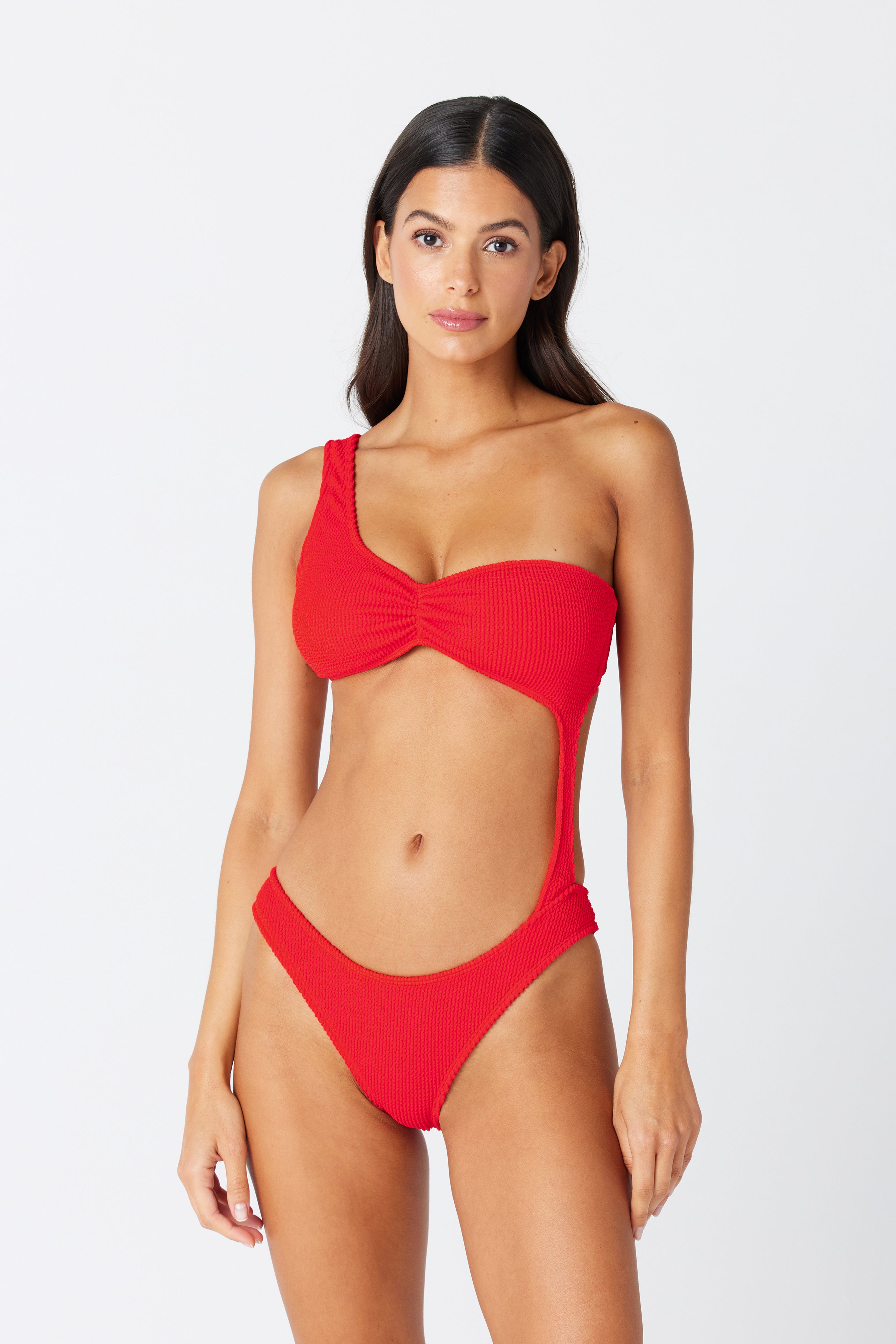 A person models the Kylie Monokini by Gigi C Bikinis against a plain white background, showcasing its red, one-shoulder cut-out style with high-cut legs, asymmetric design, open midriff, and textured crinkle jacquard fabric.