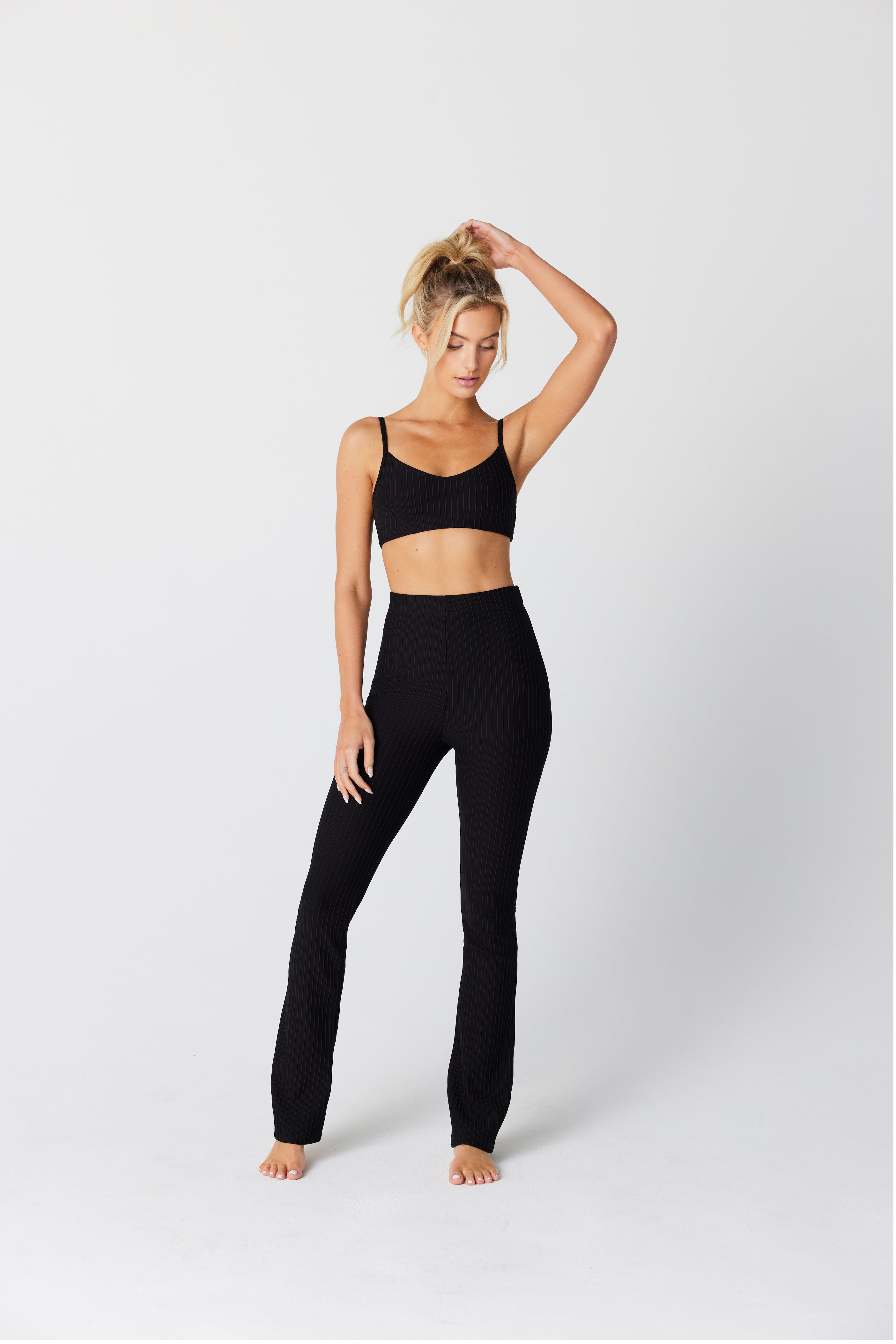 Against a plain white background, a woman confidently poses in a black ribbed crop top similar to the Lily Lounge Bra by Gigi C Lounge and high-waisted pants. Her left hand touches her hair as she stands barefoot, looking toward the camera in this exclusive fabric ensemble.