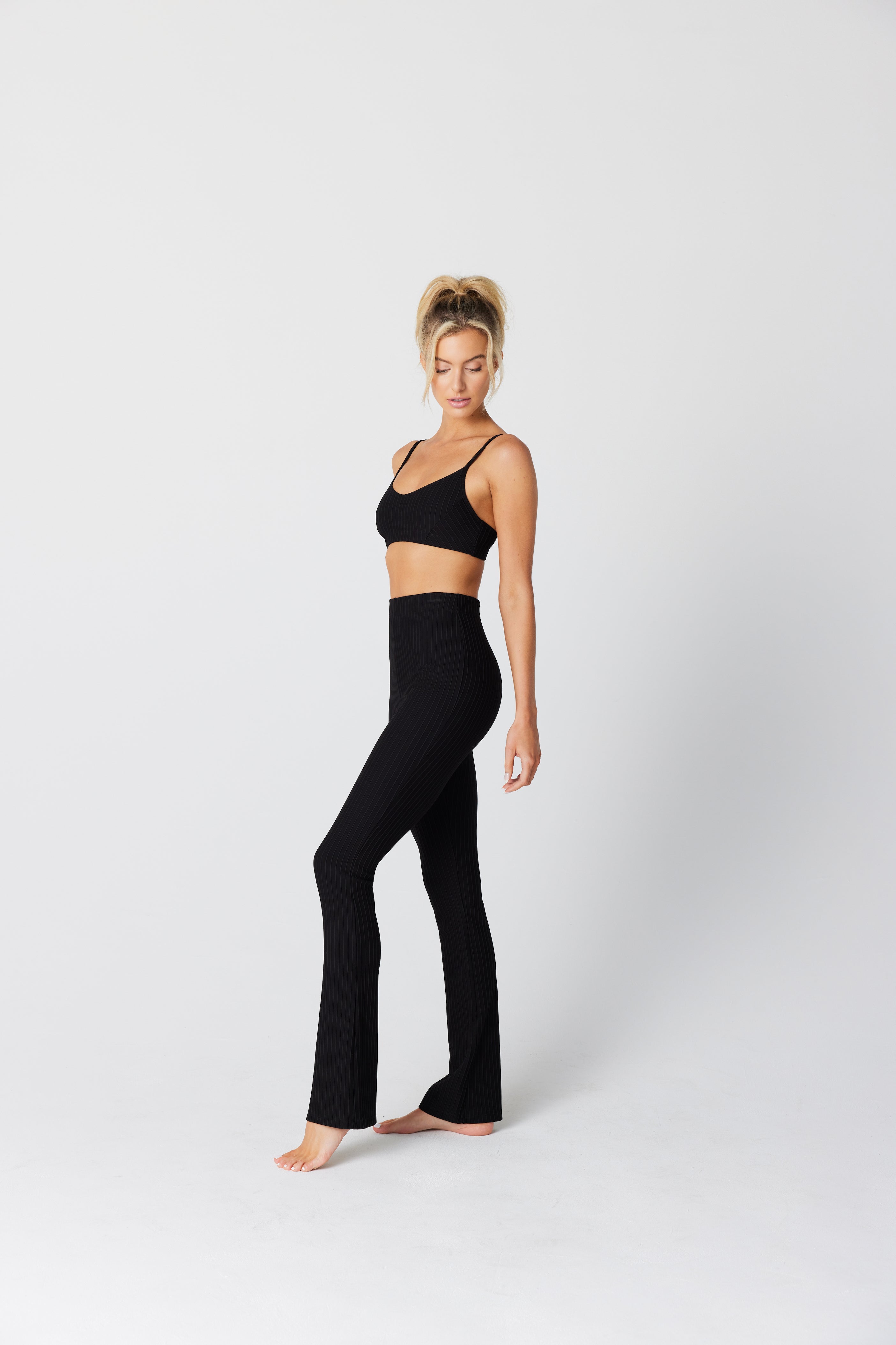 A person with a ponytail stands barefoot on a white background, wearing sleek black flared leggings and the Lily Lounge Bra by Gigi C Lounge, made from exclusive fabric. They look down to the side, embodying the essence of wireless pad-free fit comfort.