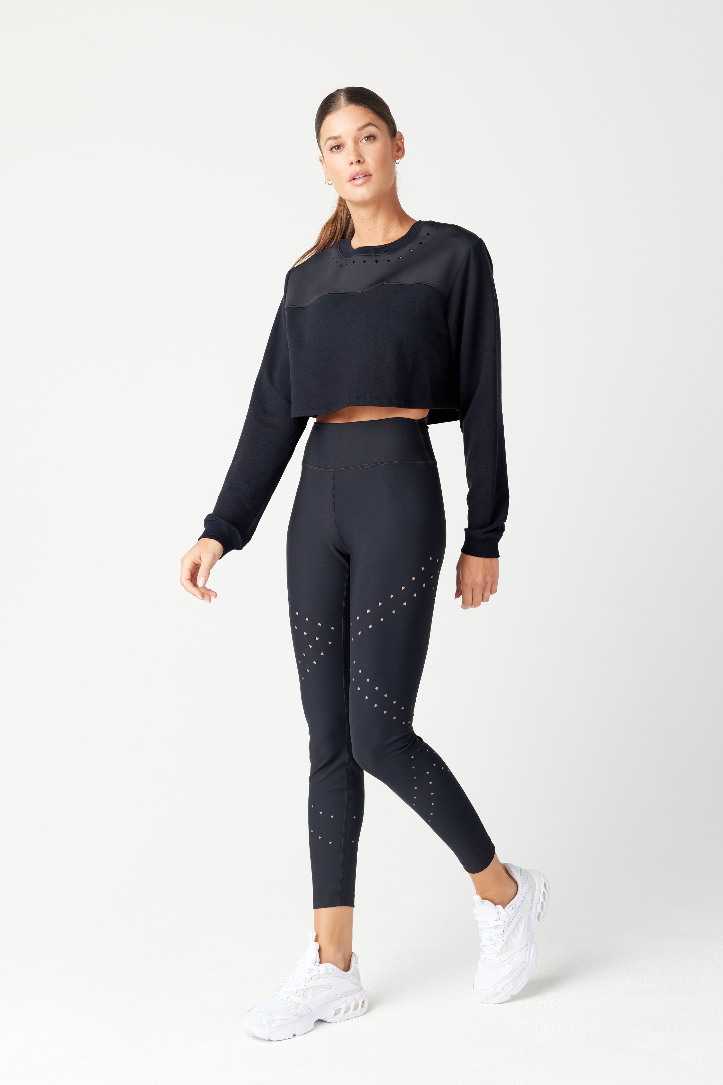 A woman stands against a plain backdrop in a Gigi C Sport Lina Pullover and matching leggings made from recycled bonded Italian jersey, showcasing triangle laser cutouts. She pairs the outfit with white sneakers and has her hair tied back, facing the camera with a neutral expression.