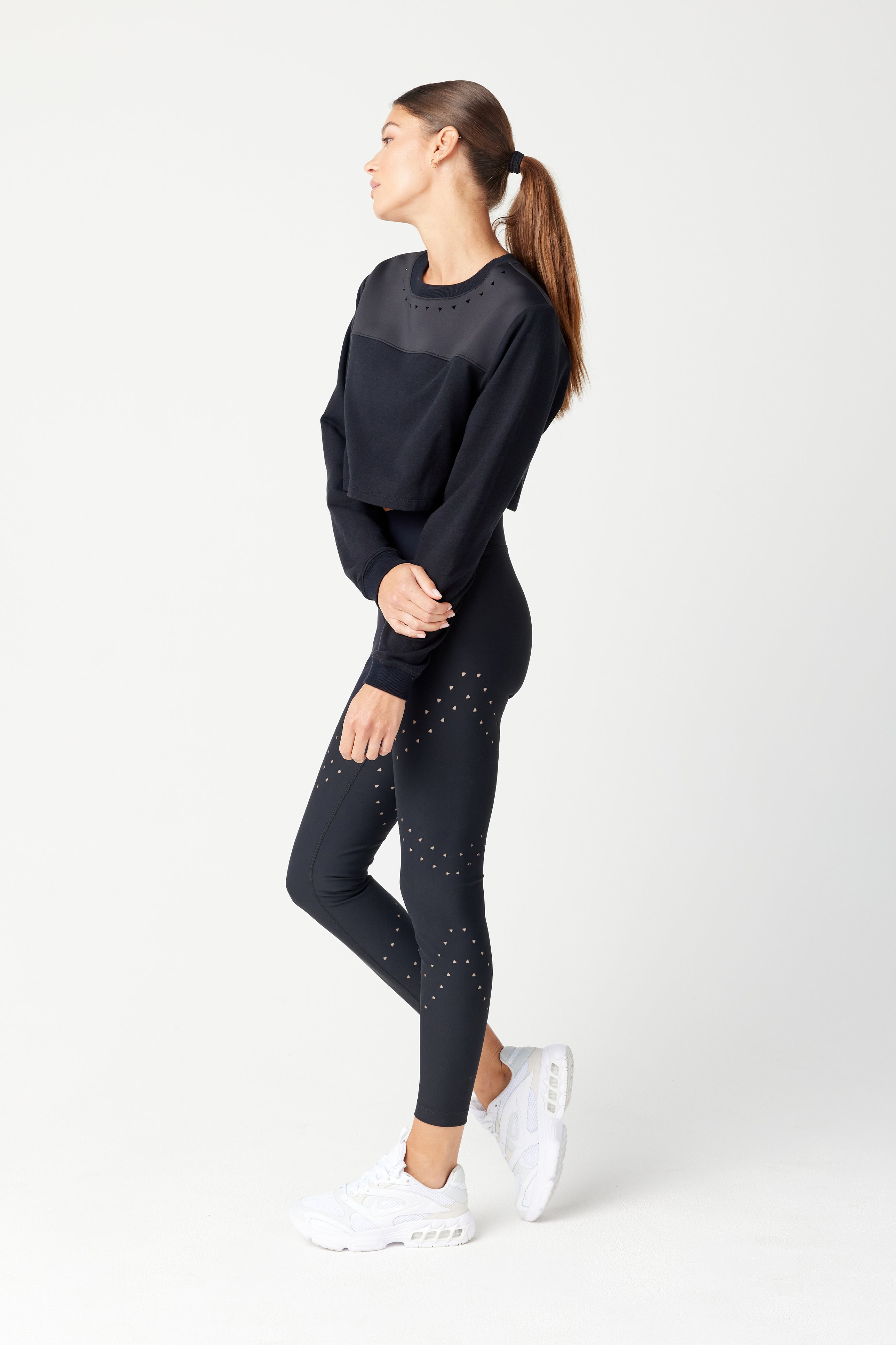 A woman wearing Gigi C Sports Lina Pullover in black activewear with triangle laser cutouts and studded details stands against a light background, looking left. Her sheer neckline adds elegance, with her hair in a ponytail. She completes her sleek look with white sneakers.