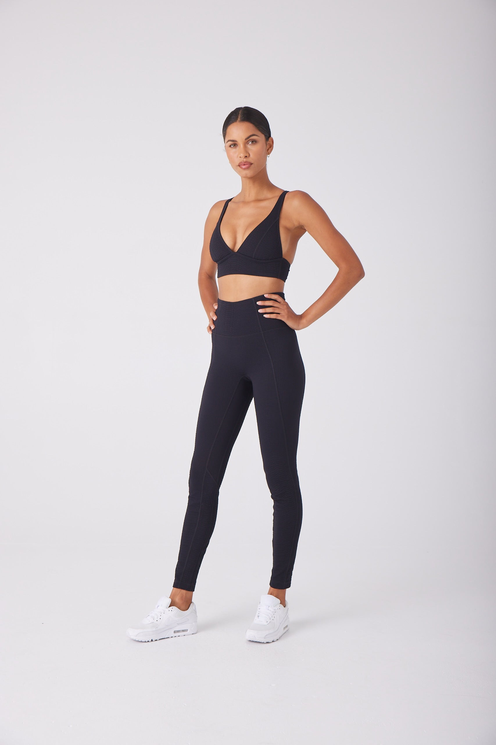 In a studio, a person confidently poses against a plain white background, wearing the Gigi C Sport Marissa Bra and matching black leggings with bi-stretch compression for medium support, paired with white sneakers. One hand rests on their hip while the other is by their side.