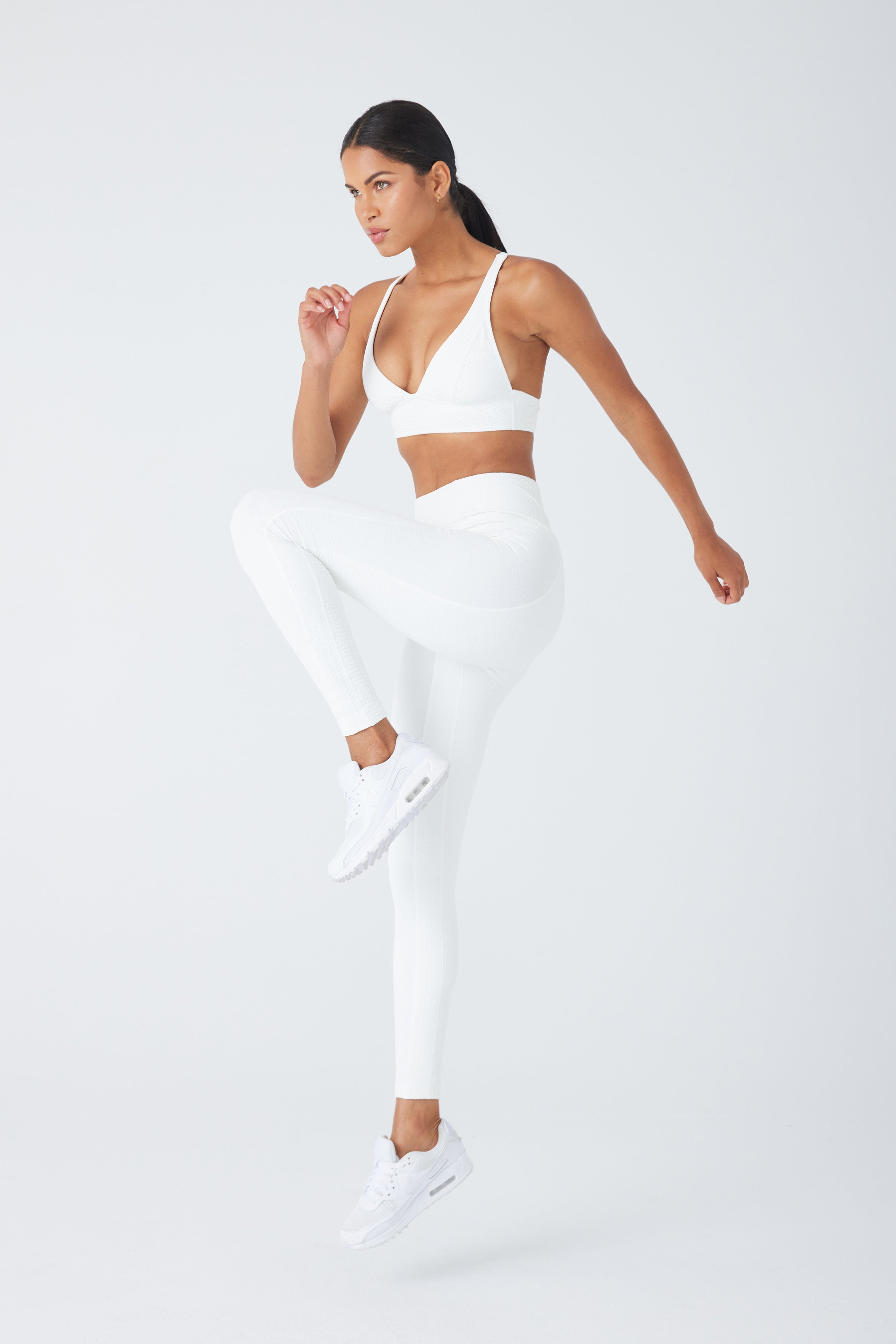 A person wearing a white Gigi C Sport Marissa Bra, faux snake leggings, and sneakers performs a jump against a plain white backdrop, one knee raised and arm bent. The pose highlights movement and athleticism, demonstrating the medium support of their outfit.