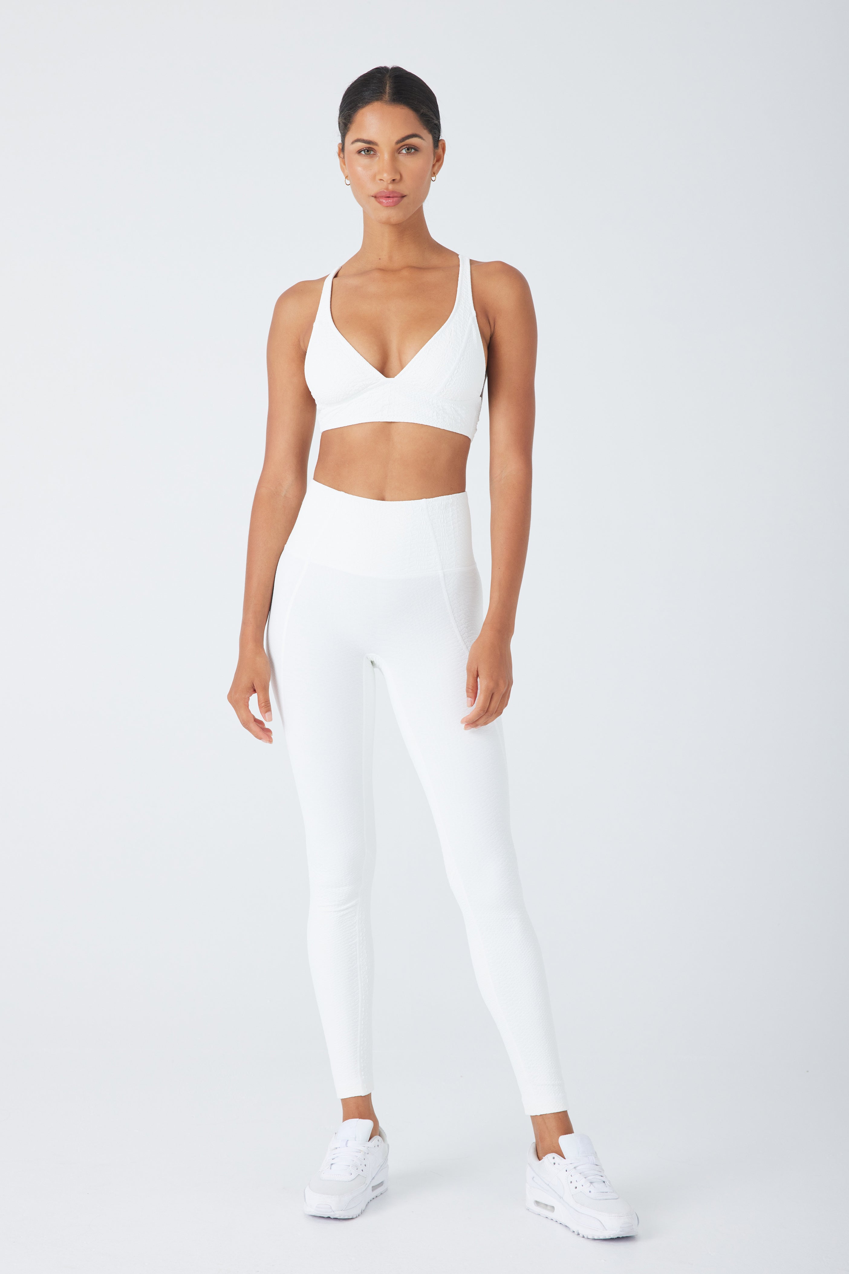 Against a light gray background, a person wears a Gigi C Sport Marissa Bra and matching high-waisted medium support leggings. They have white sneakers, hair tied back in a faux snake pattern, and stand in a relaxed pose with hands by their sides.