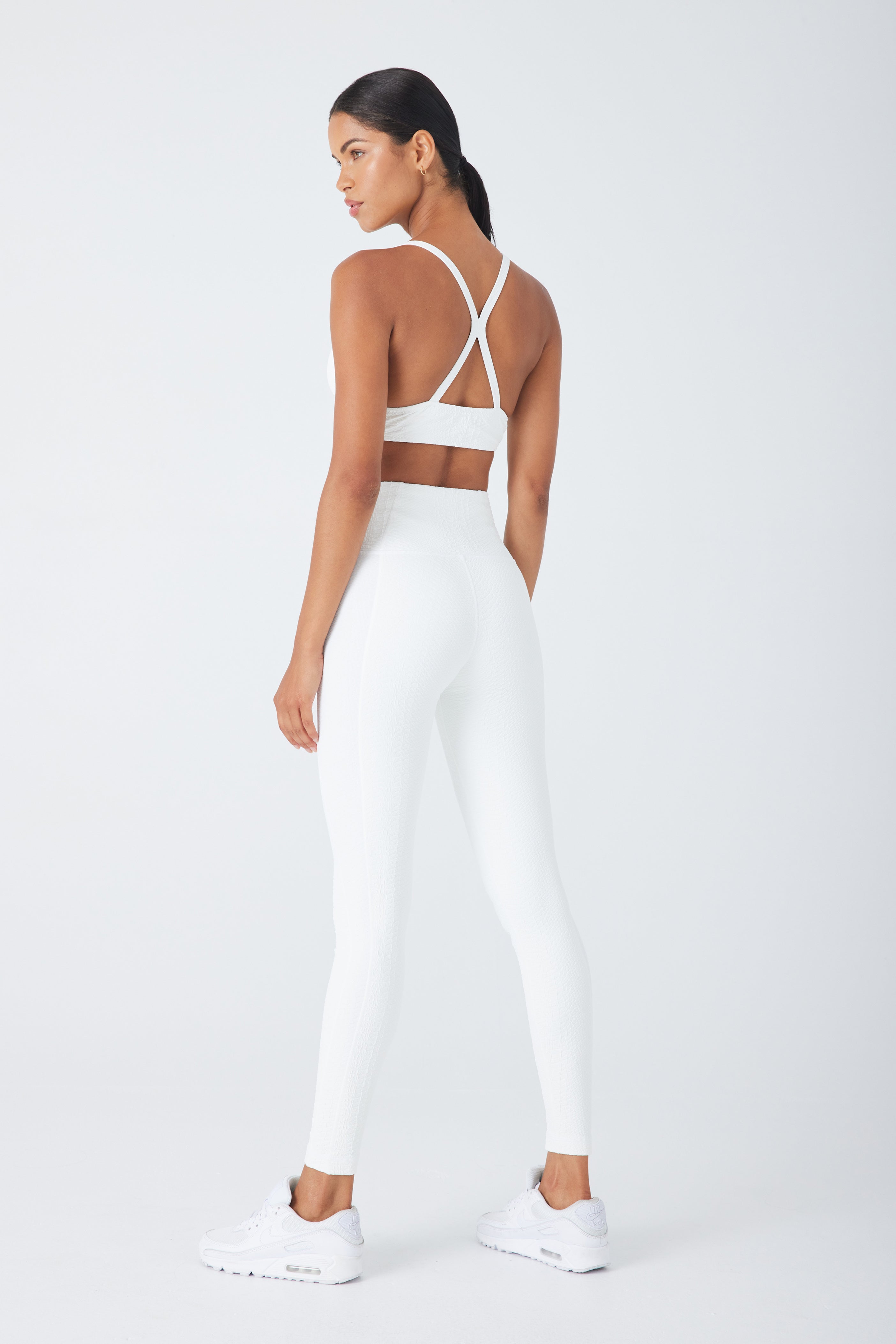 A person poses in athletic wear, looking to the side, wearing a white Gigi C Sport Marissa Bra with cross-back straps and matching high-waisted leggings for medium support. White sneakers complete the outfit on a plain, light-colored backdrop.