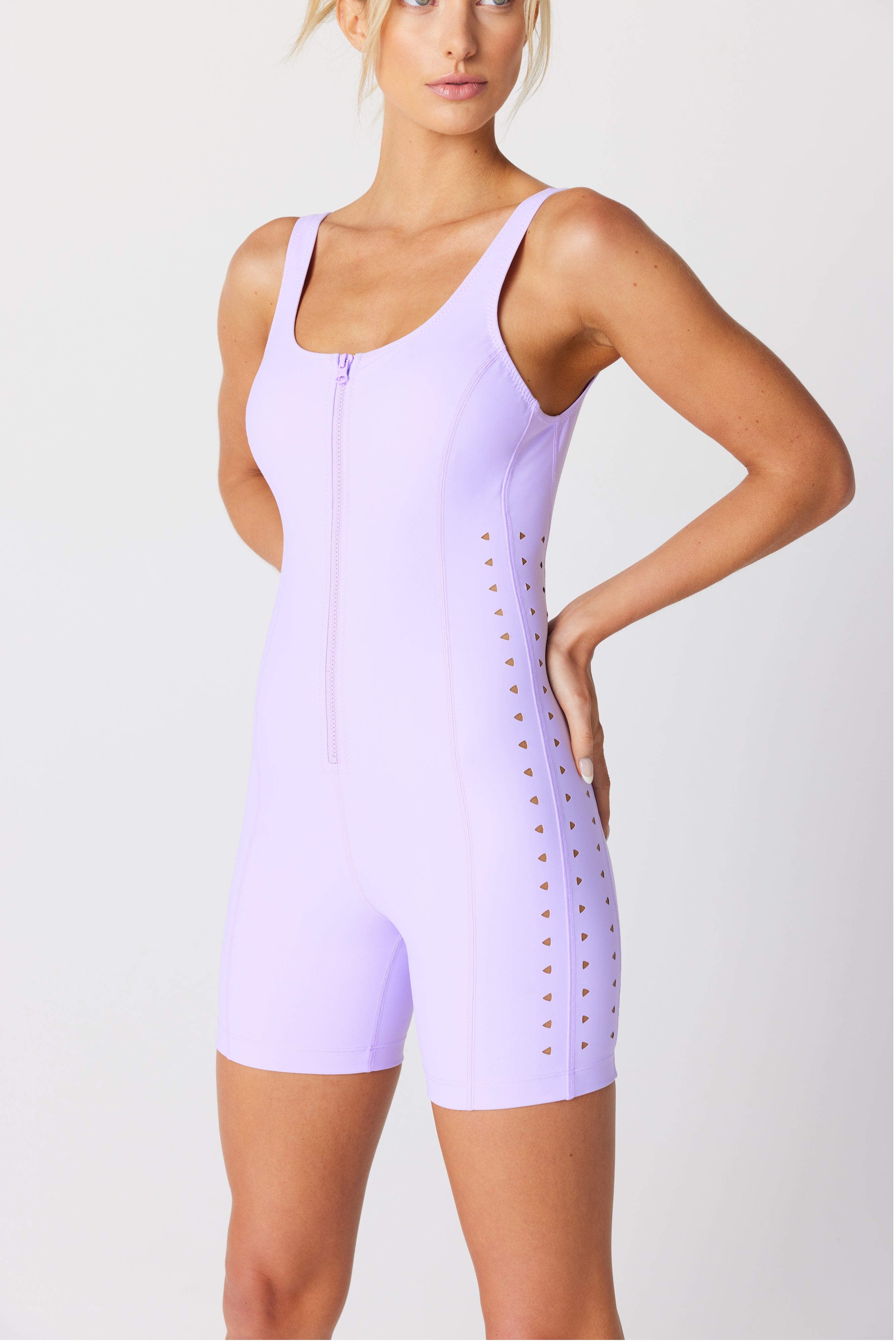 Wearing the Maxine Romper by Gigi C Sport, a person poses confidently against a neutral background. The light purple sleeveless romper features a front zipper and laser cut design, crafted from recycled Italian fabric, with their hand on their hip.