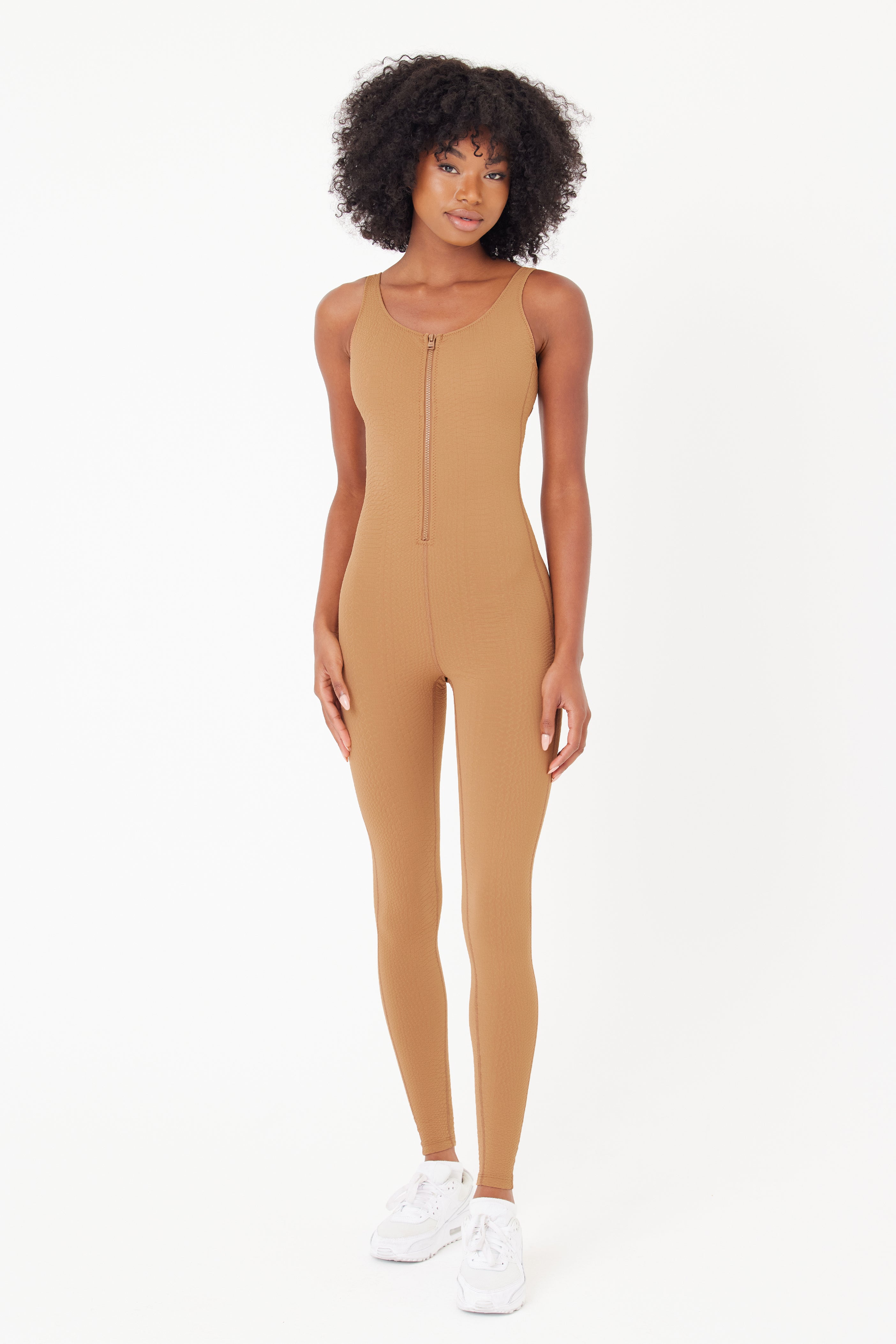 A person wears the tan, sleeveless Merit Jumpsuit from Gigi C Sport, made of waffle jacquard fabric, paired with white sneakers. Their curly hair frames a neutral expression against a plain white background.