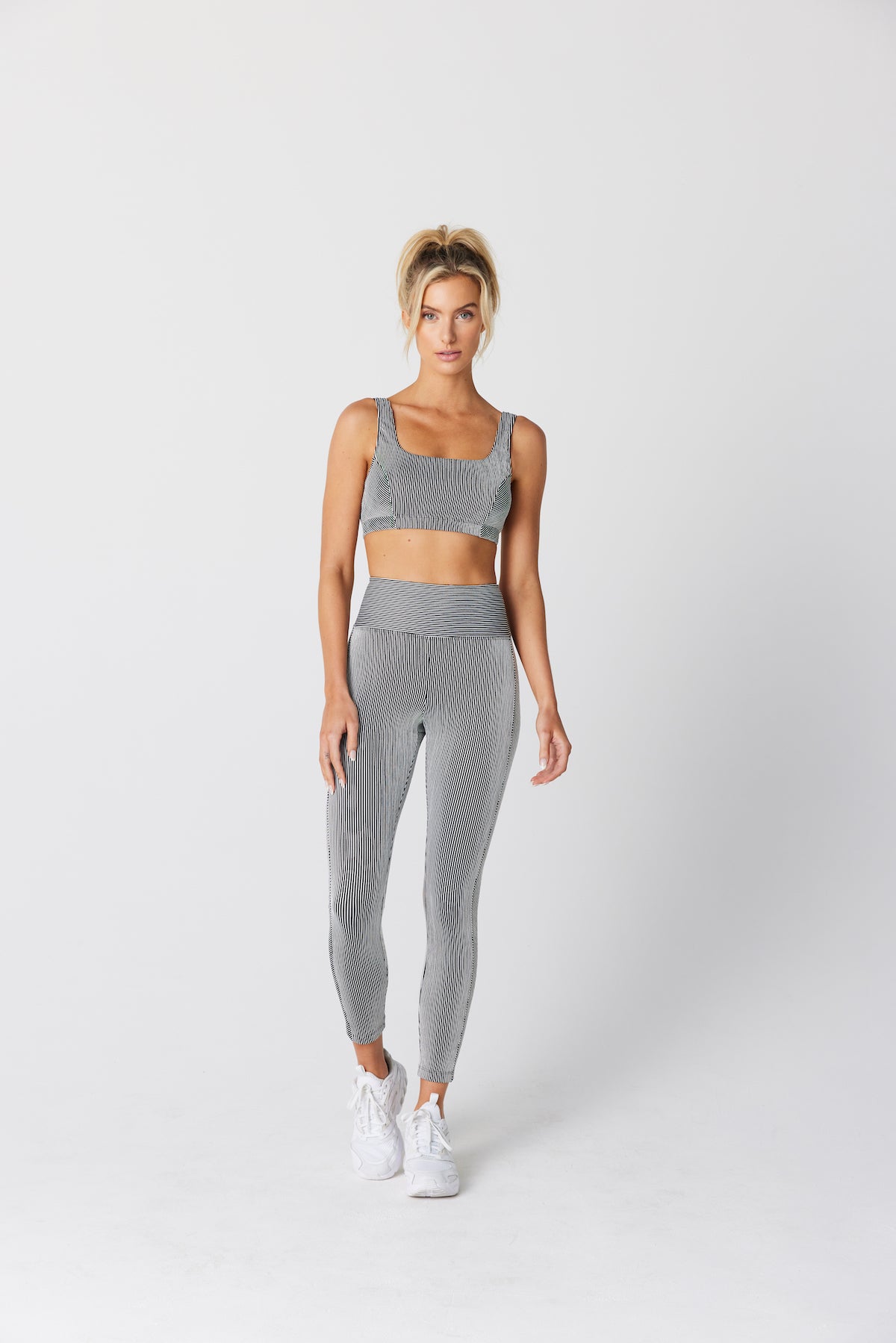 Against a plain white background, a person wears a Gigi C Sport Pamela Bra with matching gray ribbed textured fabric leggings, complemented by white sneakers and styled with a ponytail.