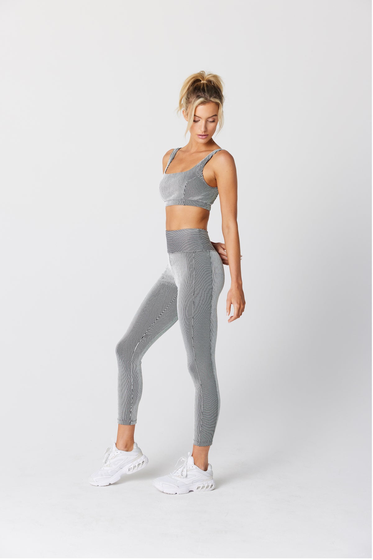 A woman stands against a light gray background, wearing the exclusive Pamela Bra and matching leggings from Gigi C Sport in a textured pattern. She pairs them with white sneakers, her blond hair in a ponytail, and strikes a relaxed pose with one foot slightly forward.