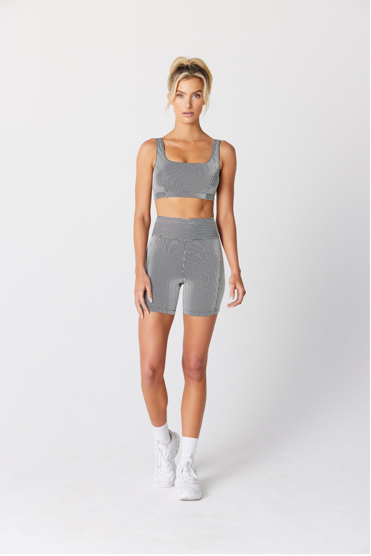 Jess Short models a gray sports bra and shorts with mini stripes from Gigi C Sport, paired with white socks and shoes. Her hair is tied up as she confidently faces the camera against a plain background.