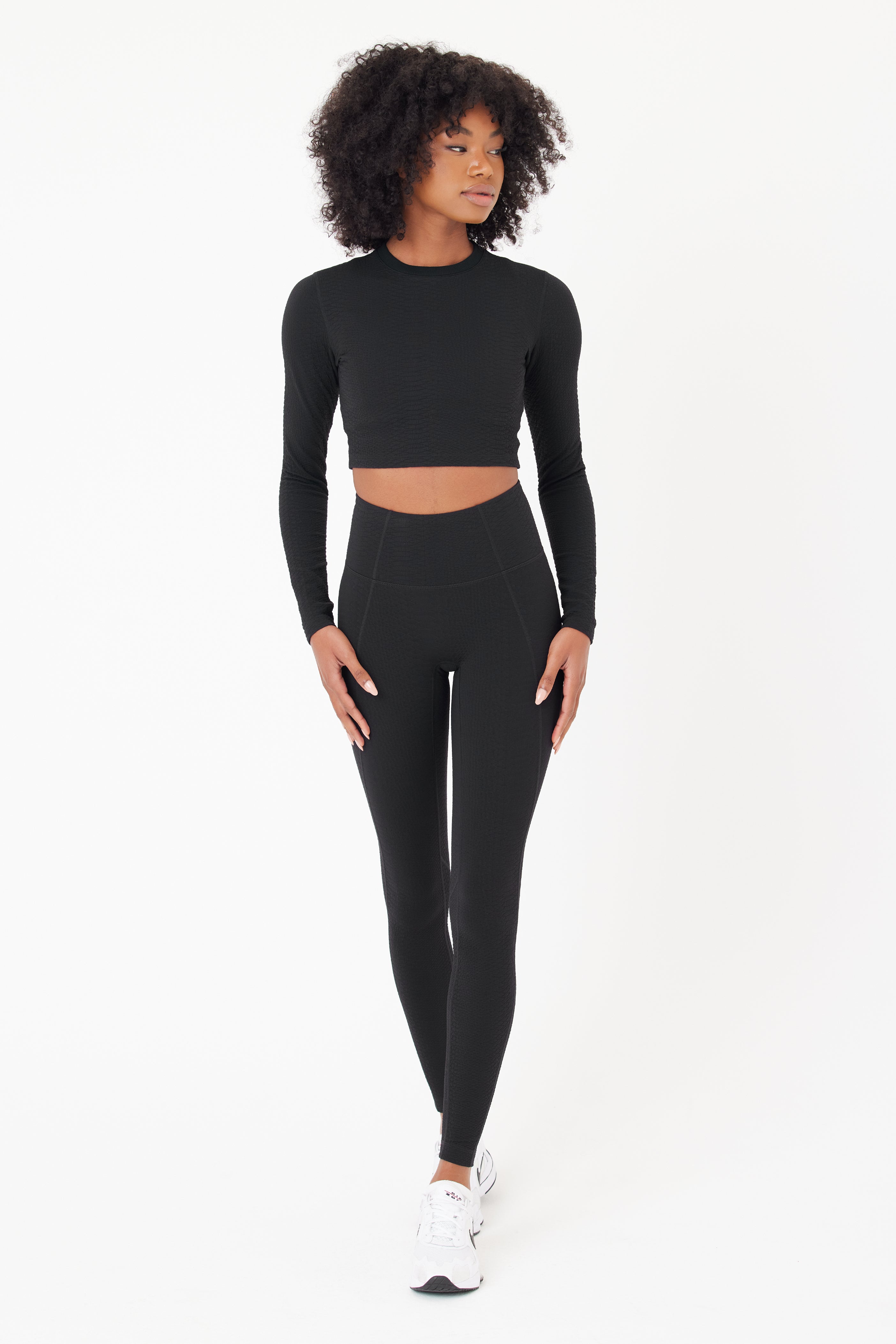 Against a plain white background, a curly-haired individual poses relaxedly, wearing Gigi C Sports sleek black Quinn Top and Ava Legging with white sneakers, blending style and comfort with hands gently resting on one knee.