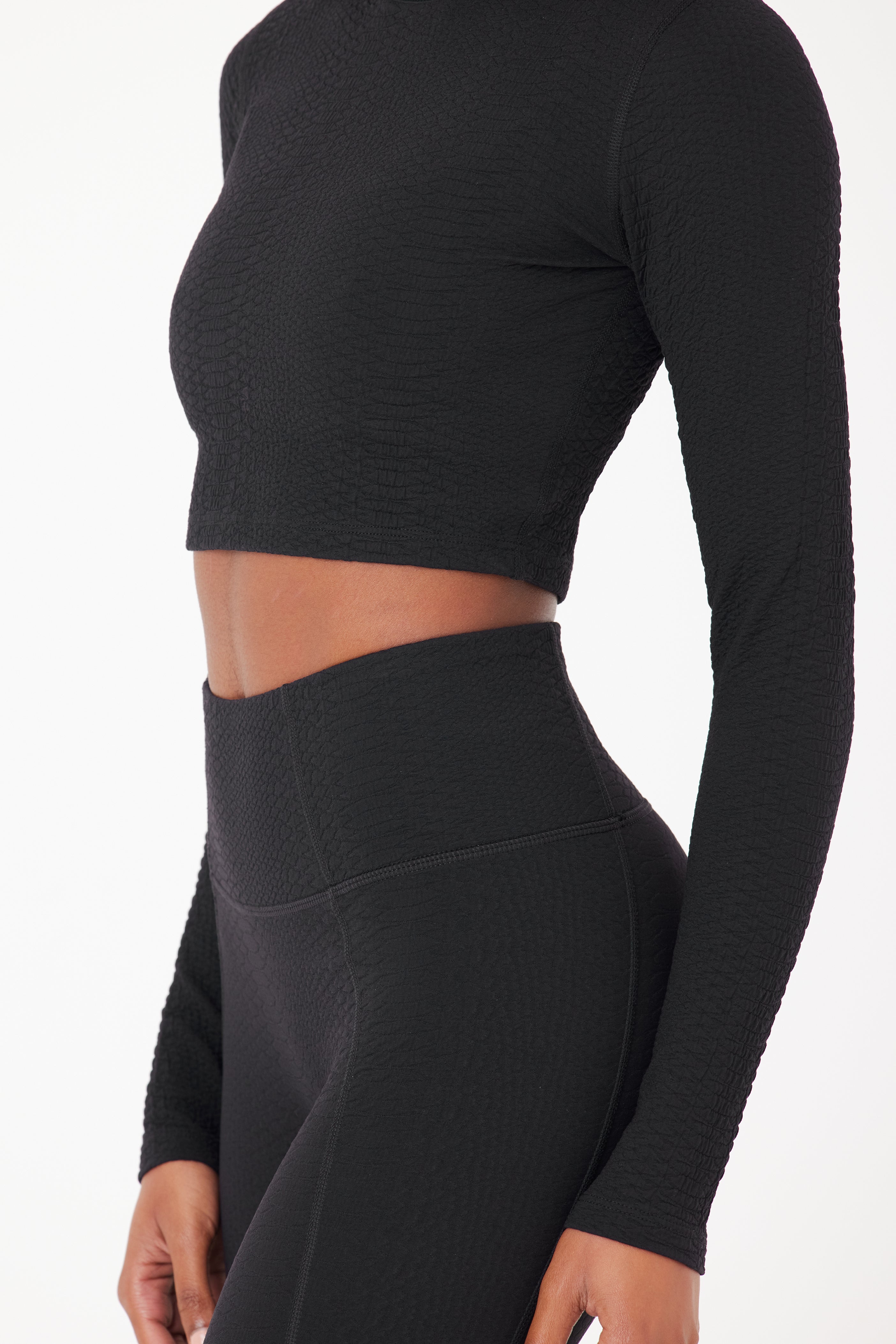 A person wears a textured black long-sleeve Quinn Top by Gigi C Sport and high-waisted Ava Legging, showcasing a form-fitting style with a minimalistic faux snake design against a plain white background.