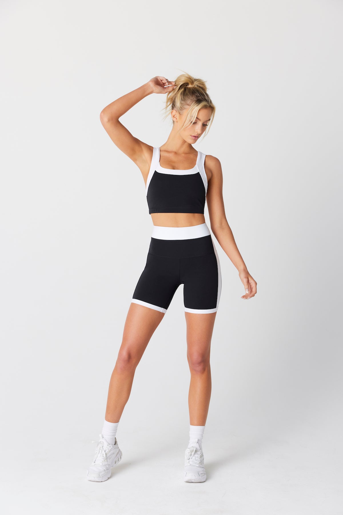 Natasha Short poses elegantly in a Nadia crop top and Natasha Bike Short by Gigi C Sport. Against a plain backdrop, her ponytail sways as she lifts an arm to highlight the quick-drying fabric and four-way stretch shorts, complemented with sleek white socks and sneakers.