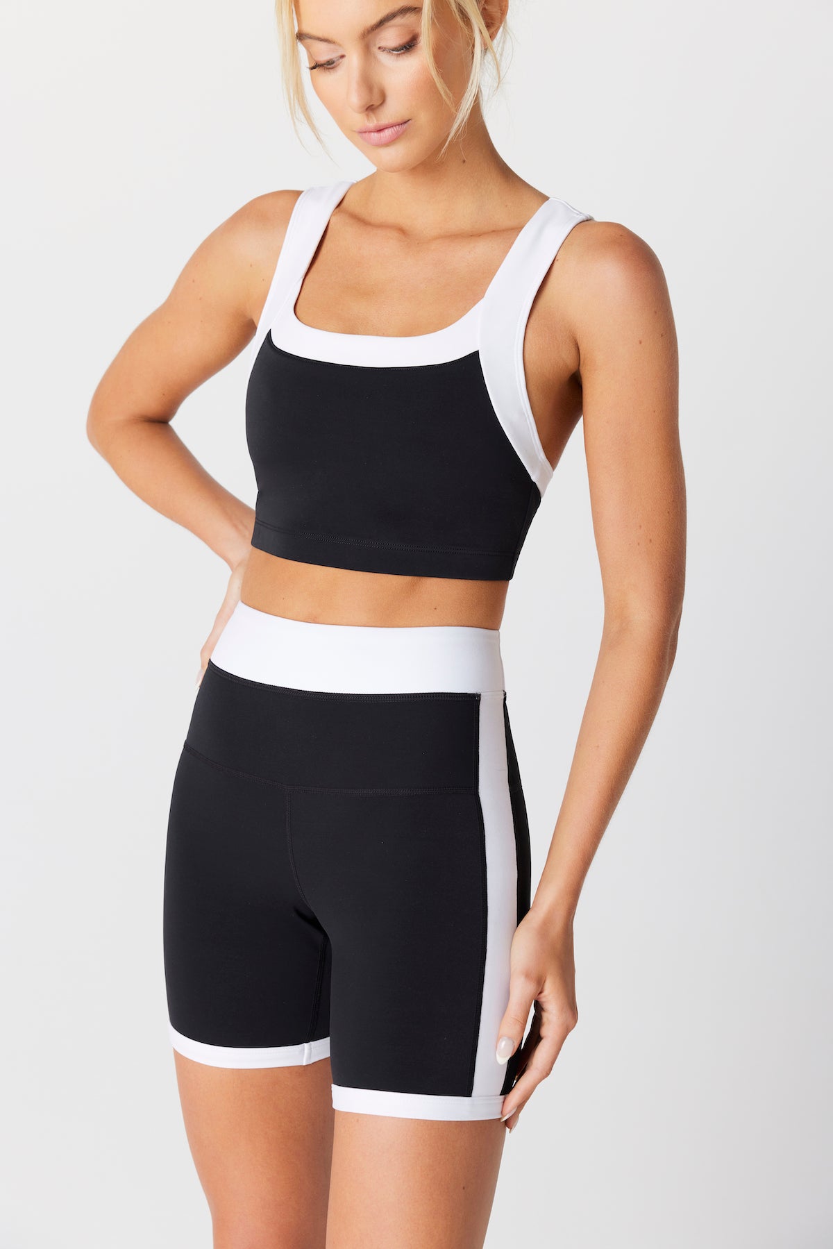 Wearing the Gigi C Sport Natasha Bike Short collection in black and white, the woman dons a sports bra with high-waisted shorts. Crafted from quick-drying, four-way stretch fabric for comfort, she gazes thoughtfully downward with her hand on her hip against a plain backdrop.