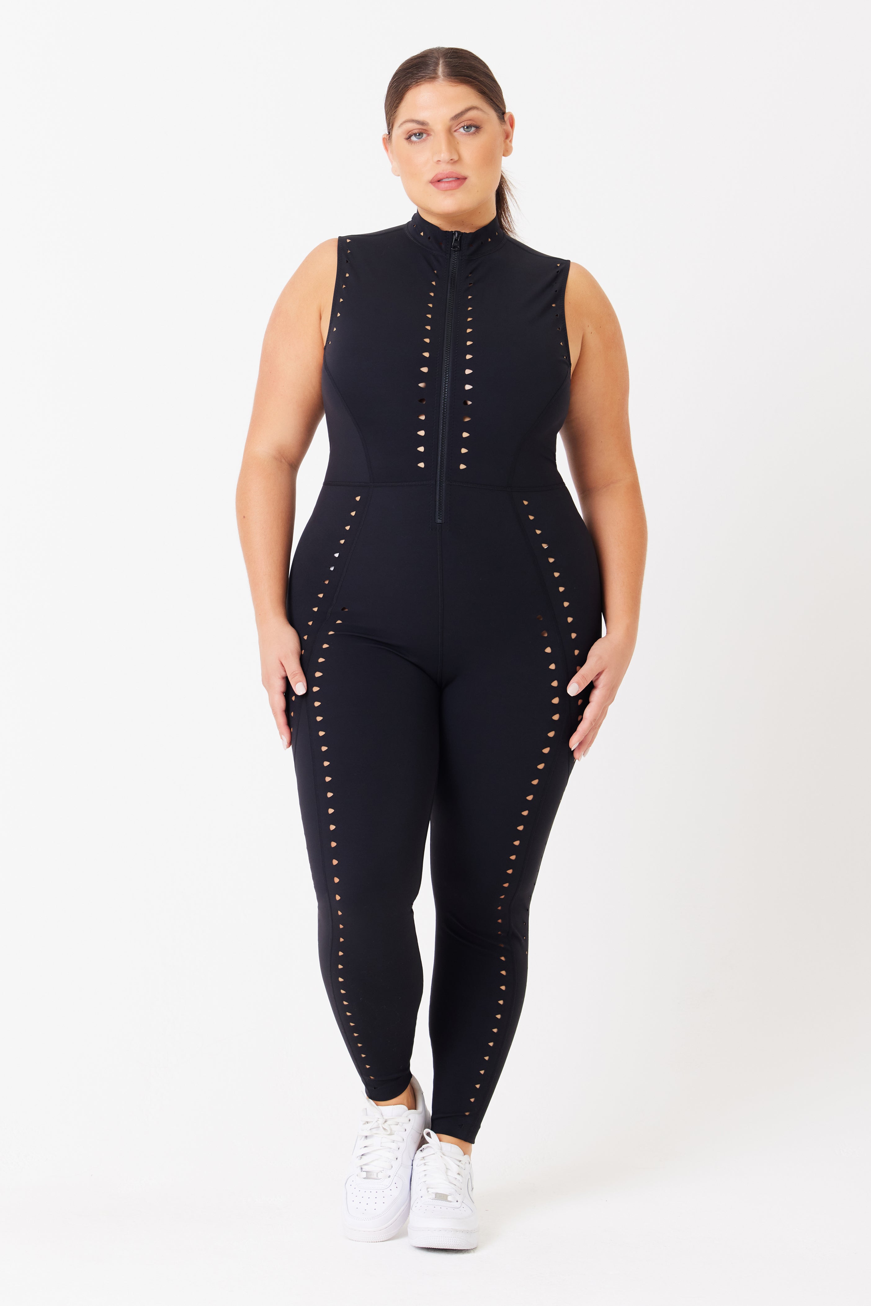 A person is styled in a fitted, sleeveless Sarah Jumpsuit by Gigi C Sport, featuring decorative studs and laser cutout designs along the front and sides. They wear white sneakers and stand against a plain white background.