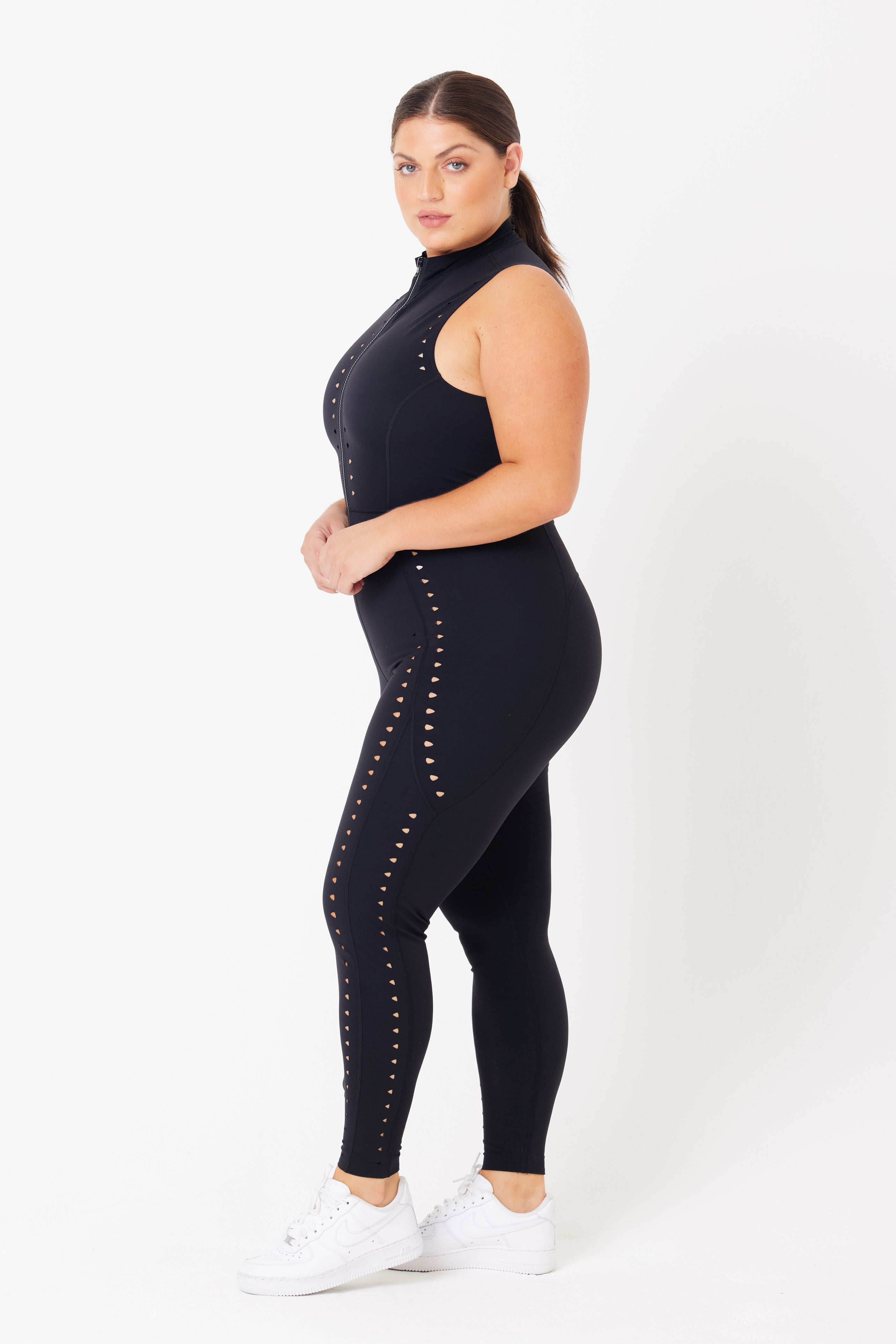 In a sleek LYCRA Sarah Jumpsuit by Gigi C Sport with intricate laser cut designs on the sides, a person stands sideways. Their ponytail complements the outfit, and white sneakers provide contrast against the plain white background.