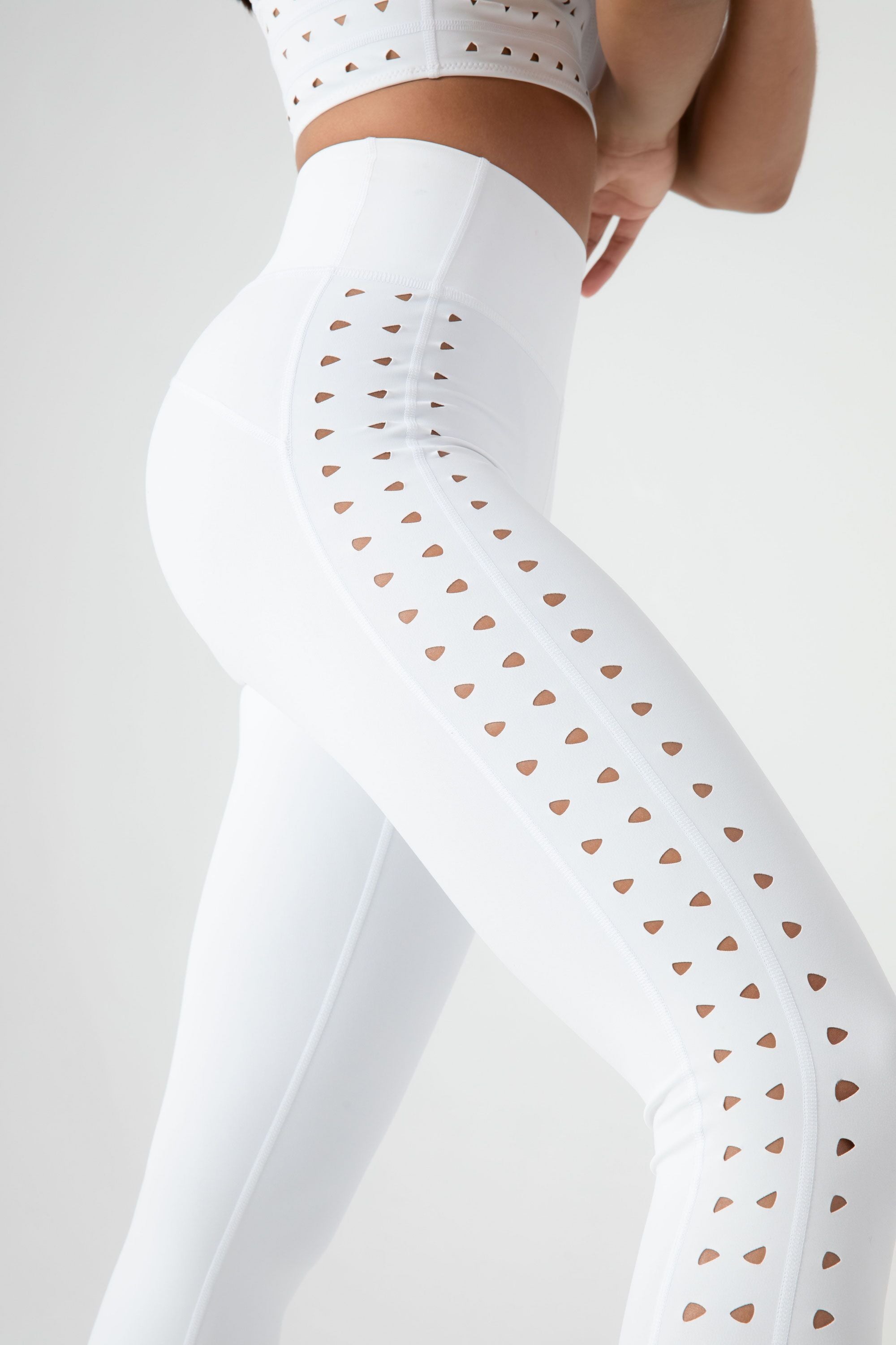 A person is wearing a stylish white athletic outfit featuring the Katrina Legging by Gigi C Sport, which has sleek laser cutouts. This design combines chic style with comfortable compression fabric against a plain background.